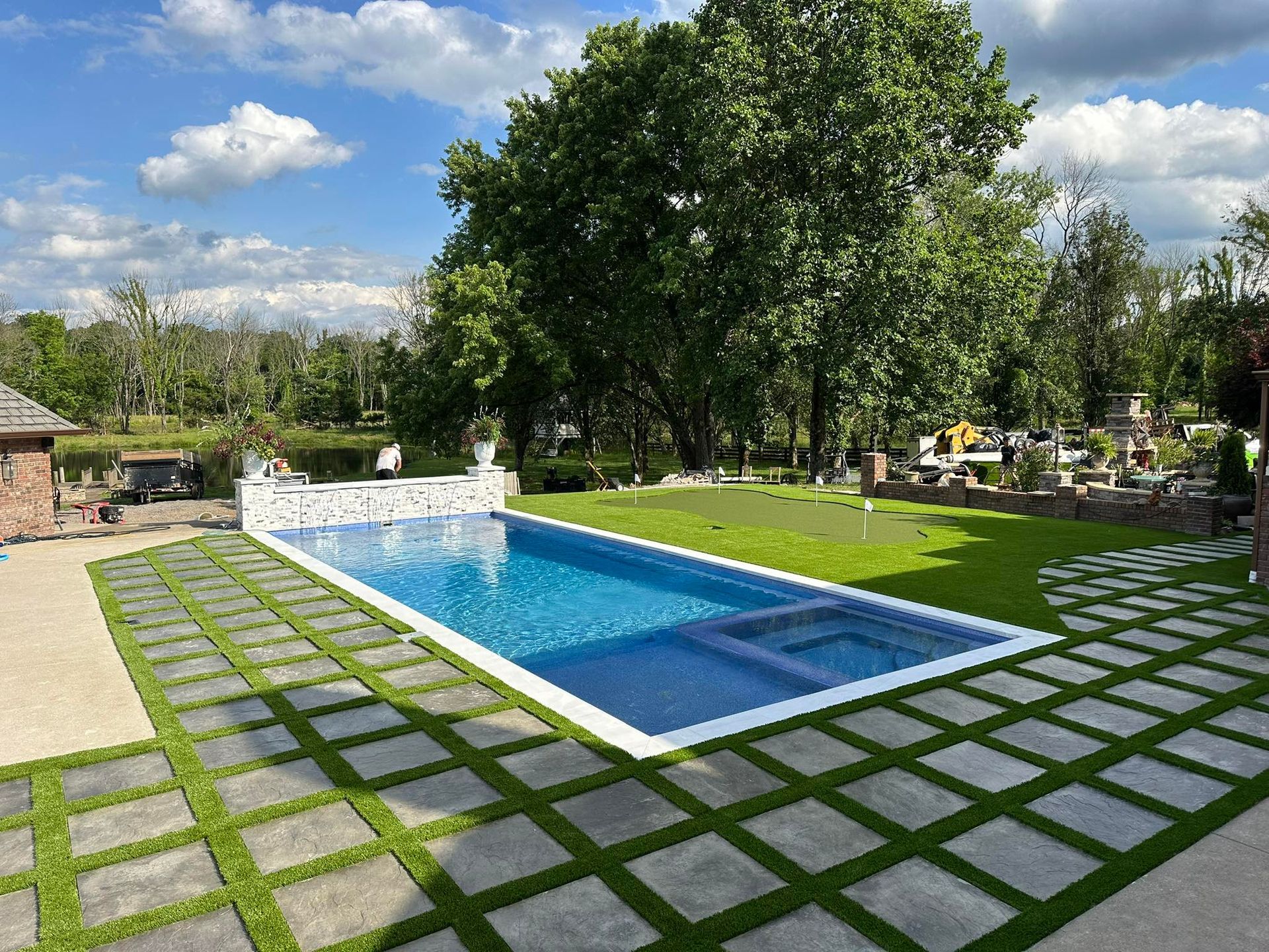 Swimming pool surrounded by green grass, stone pavers, and trees on a sunny day.