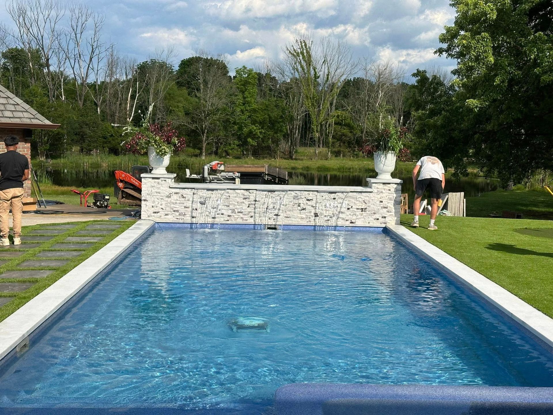 A rectangular pool with blue tiles, two people in the yard, and trees in the background under a cloudy sky.