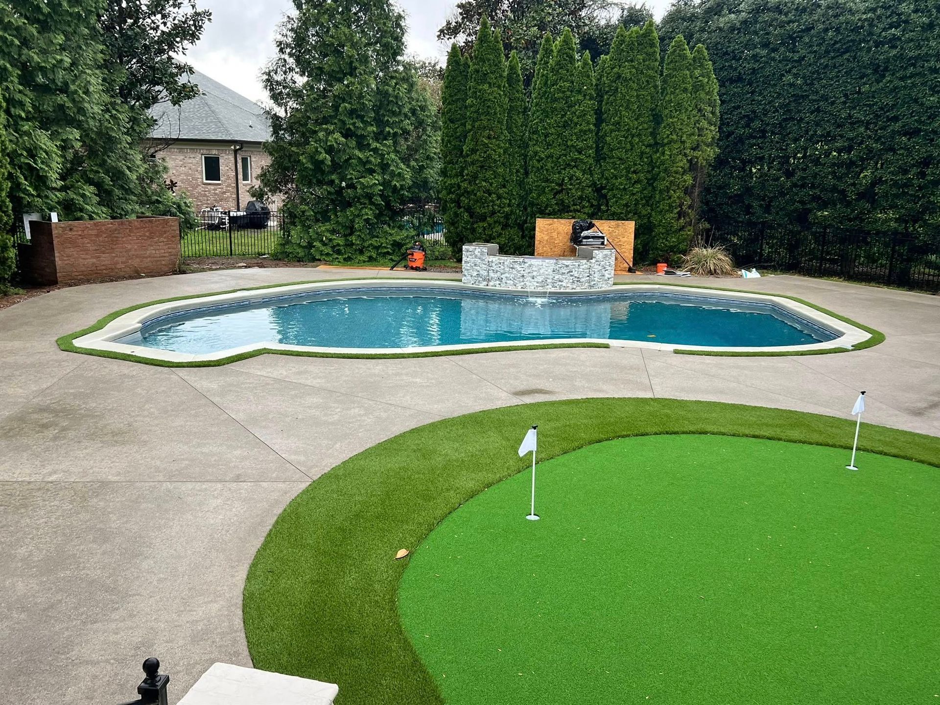 Backyard with pool and putting green surrounded by greenery.