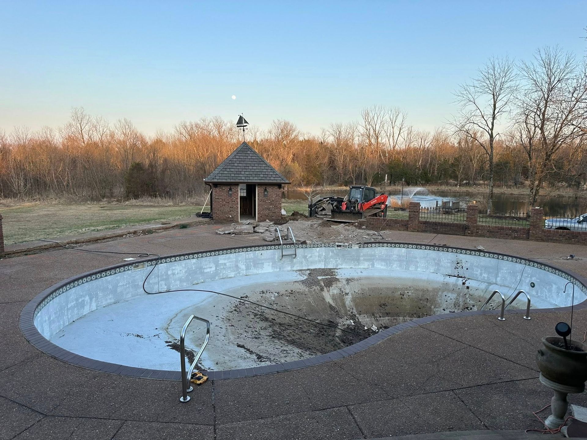 An empty, oval-shaped pool under construction with a small brick building nearby and trees in the background.