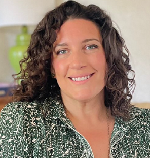 Woman with curly brown hair smiles at the camera, wearing a green patterned shirt, in a room.