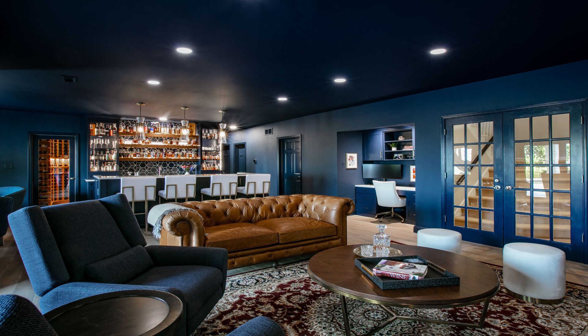 A luxurious, blue-walled lounge with a bar, tan leather sofa, and office space, lit by recessed ceiling lights.