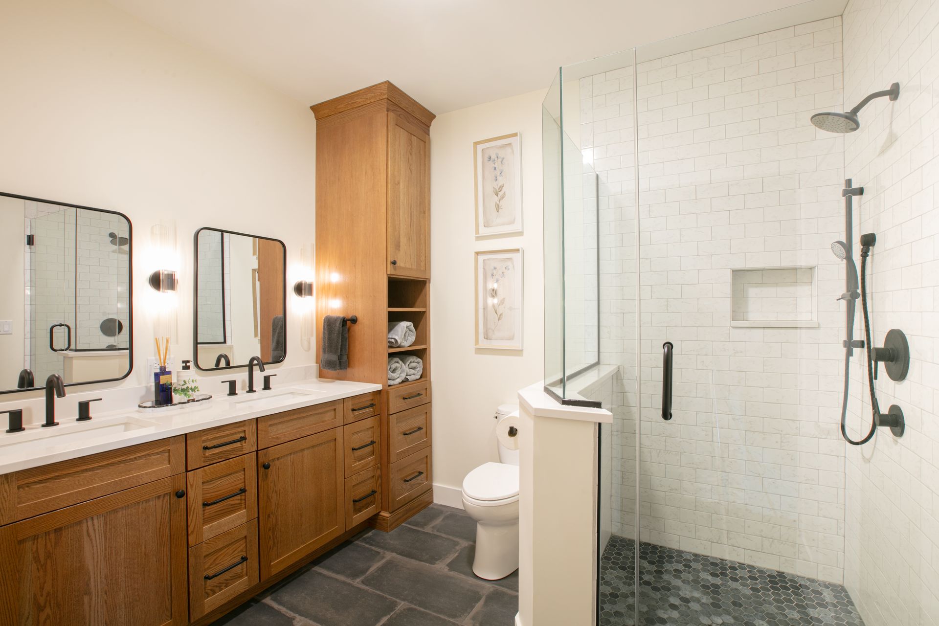 Bathroom with wood vanity, black fixtures, tiled shower, and dark stone floor.