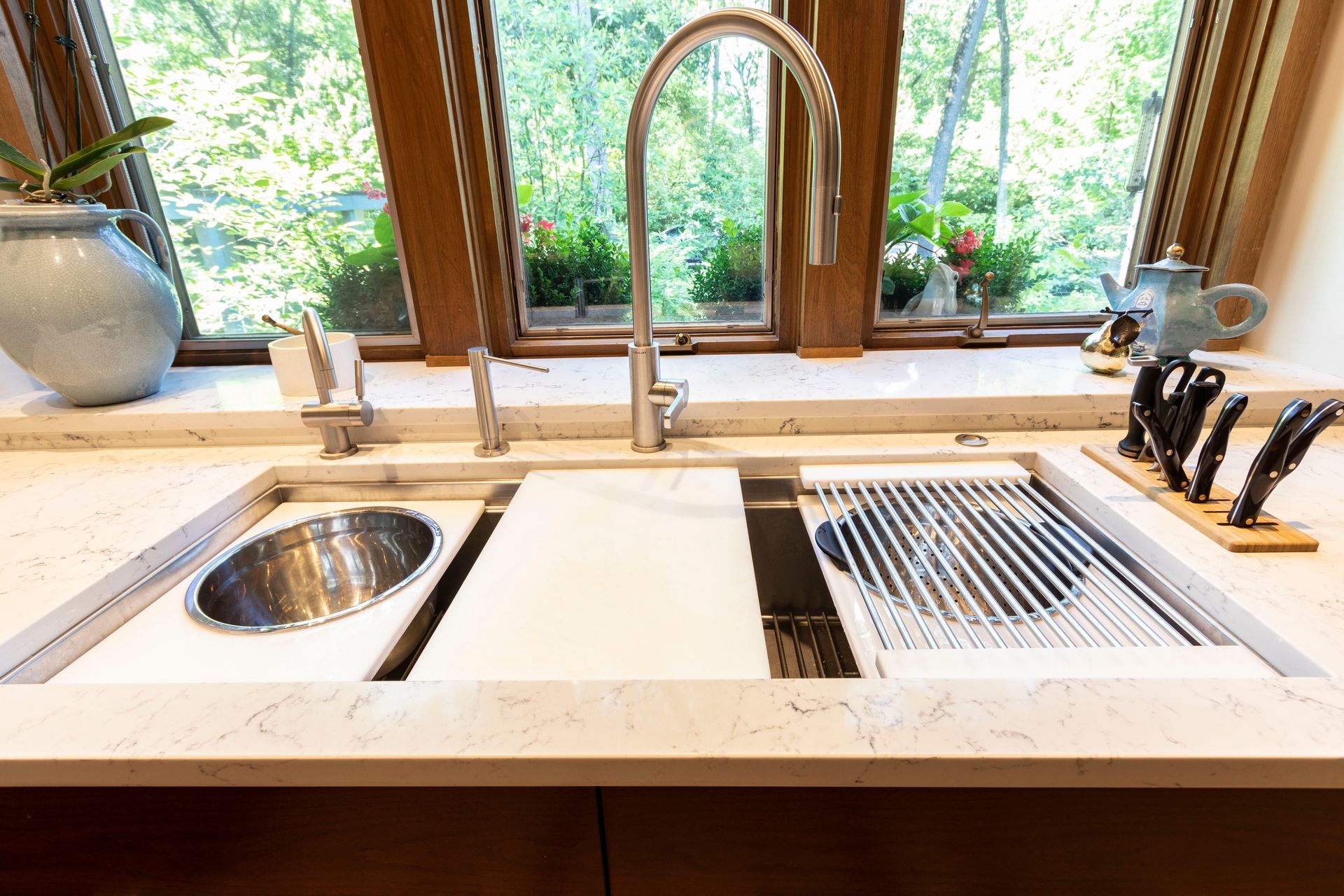 Kitchen sink with integrated accessories: colander, cutting board, and prep bowl. Window in the background.