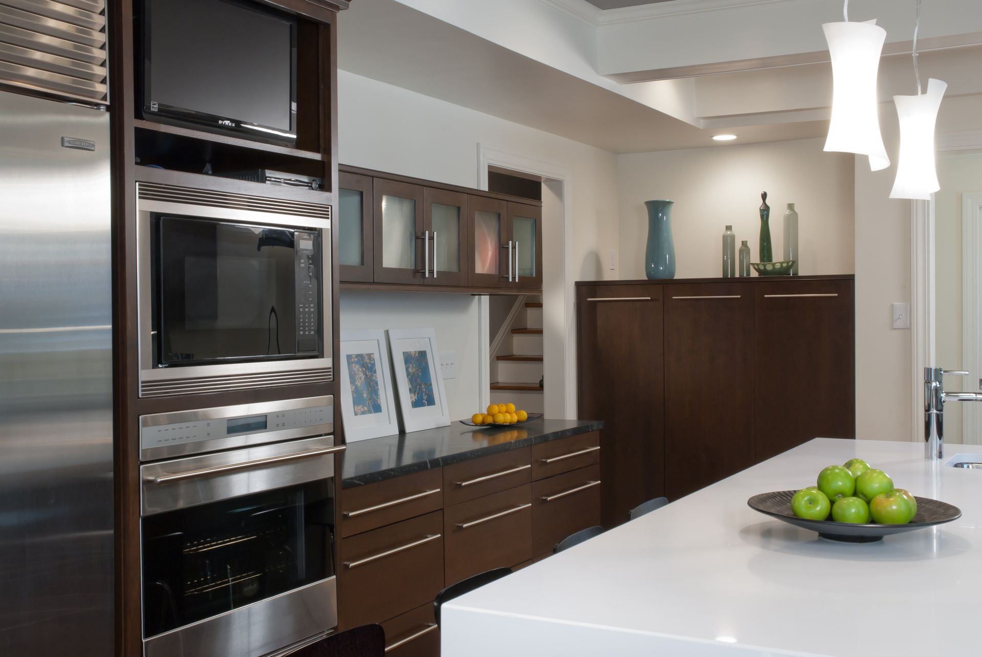 Modern kitchen with dark wood cabinetry, stainless steel appliances, and a white countertop with a bowl of green apples.