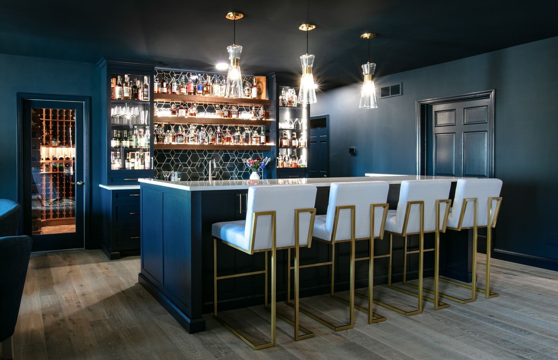 Dark blue home bar with gold accents, stocked shelves, bar stools, and pendant lights.