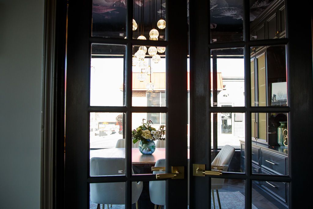 Black French doors with panes, leading to a room with table, chairs, flowers, and a chandelier.