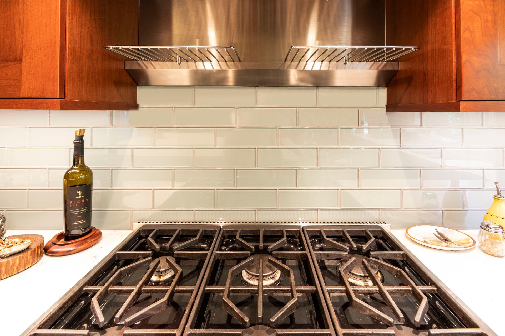 Close-up of a kitchen stovetop with range hood, light-colored backsplash, and wood cabinetry.