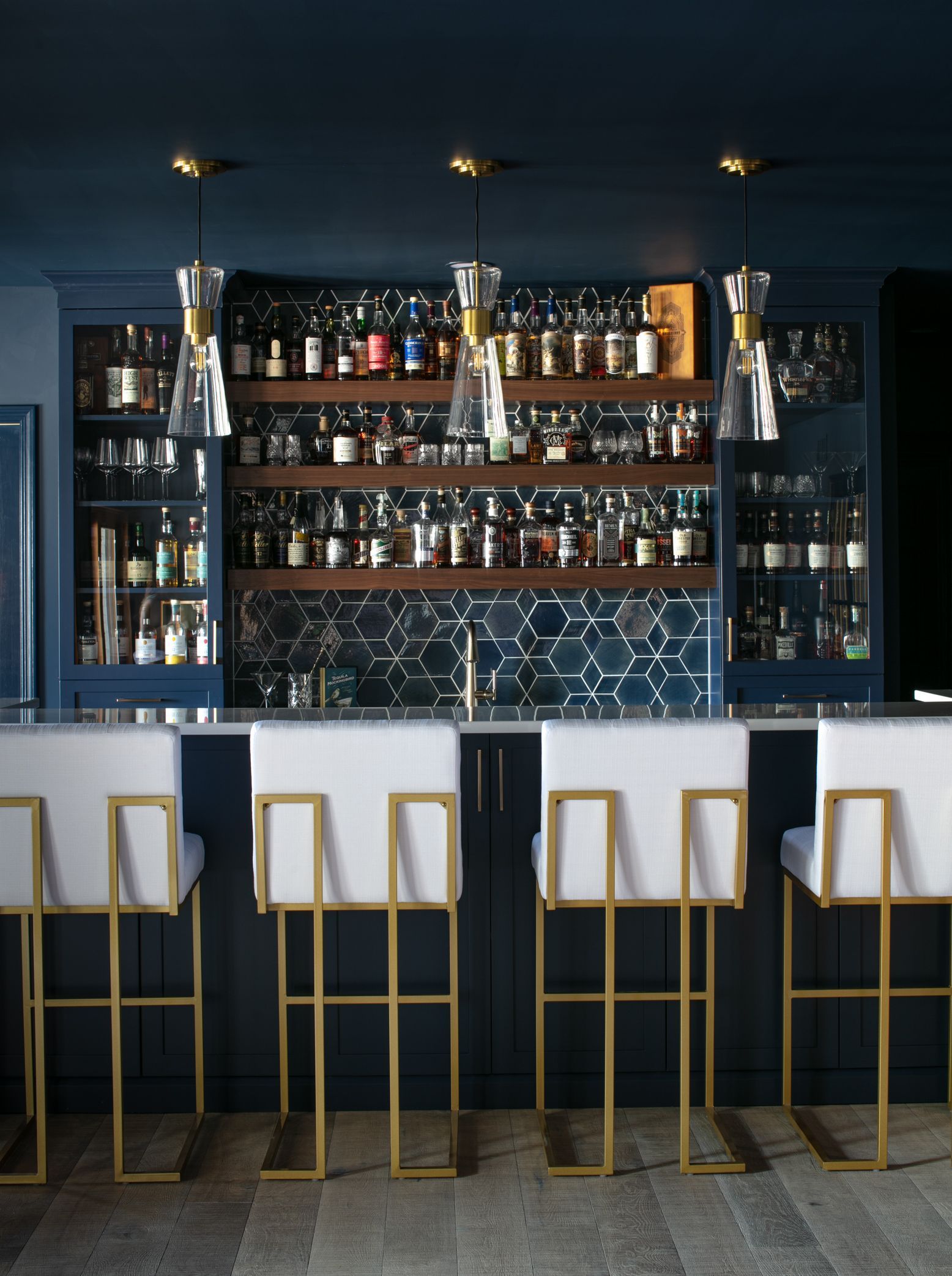 Stylish bar with navy blue cabinetry, marble-top counter, and gold-framed bar stools. Bottles on shelves.