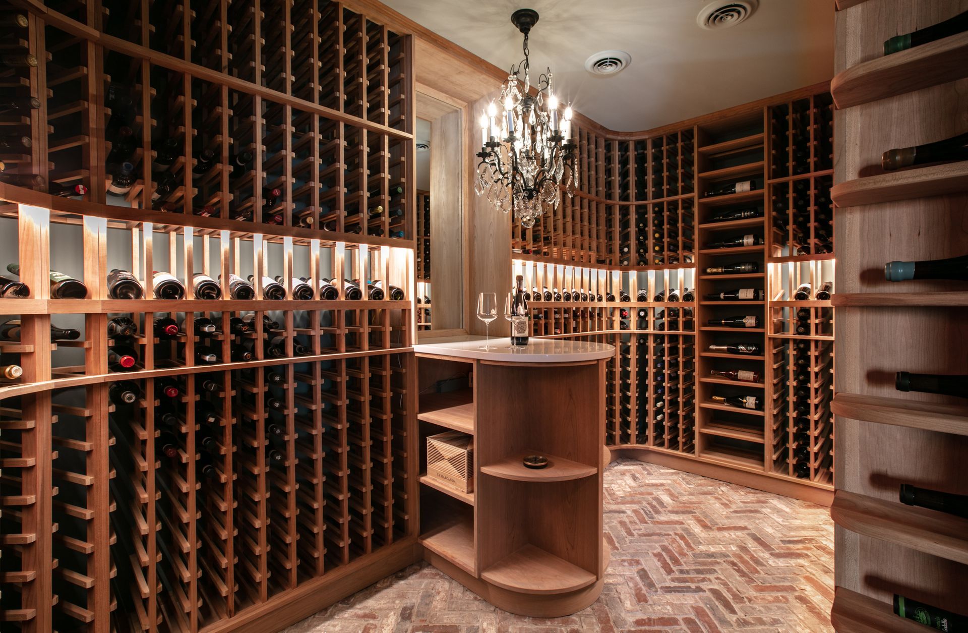 Wine cellar with wood shelving filled with bottles, herringbone brick floor, and a small tasting table.