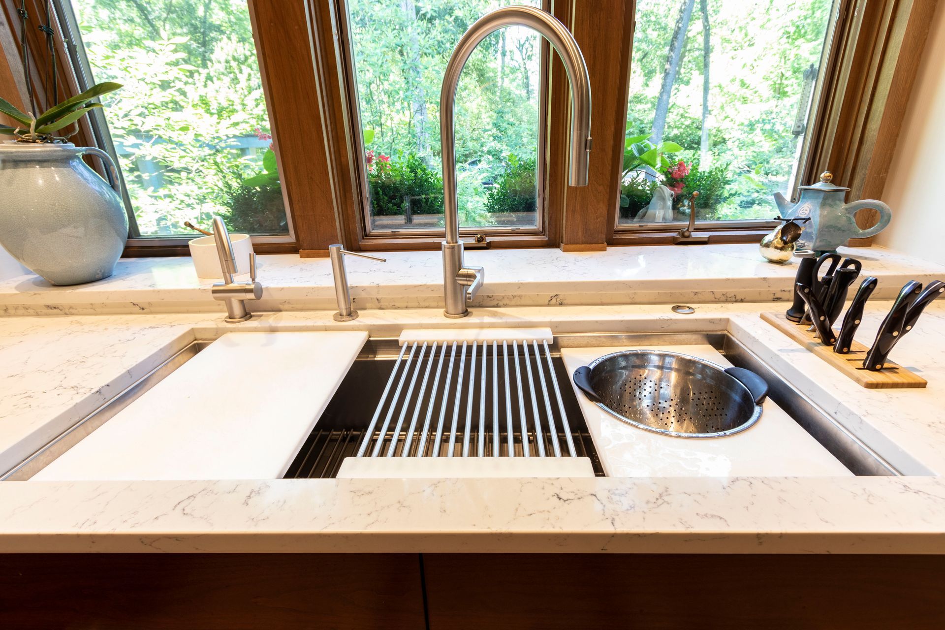 Kitchen sink with faucet, stainless steel grid, and cutting board. A window with trees in the background.