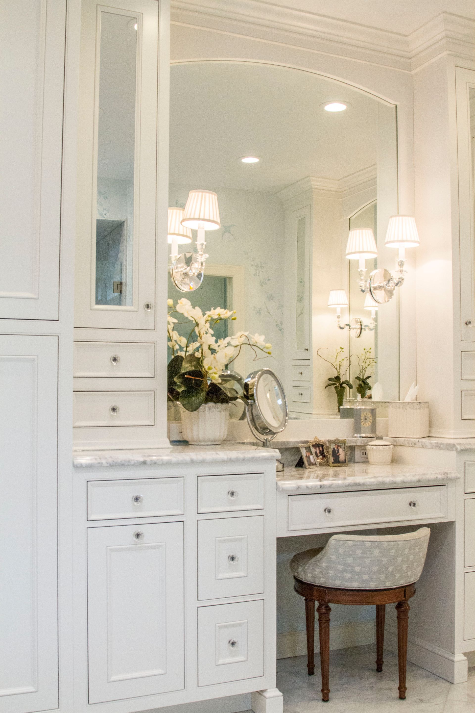 White vanity with marble countertop and large mirror; floral arrangement; patterned chair.