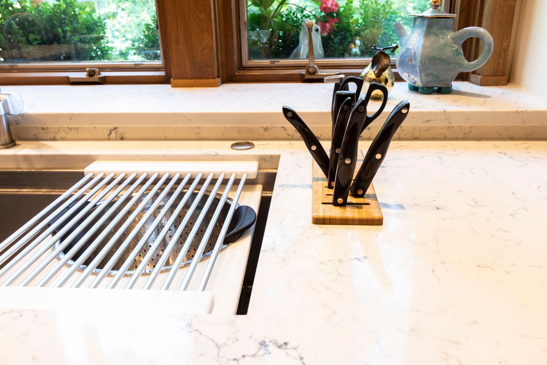 Kitchen sink with a rack, knives, and a window in the background. White countertop.