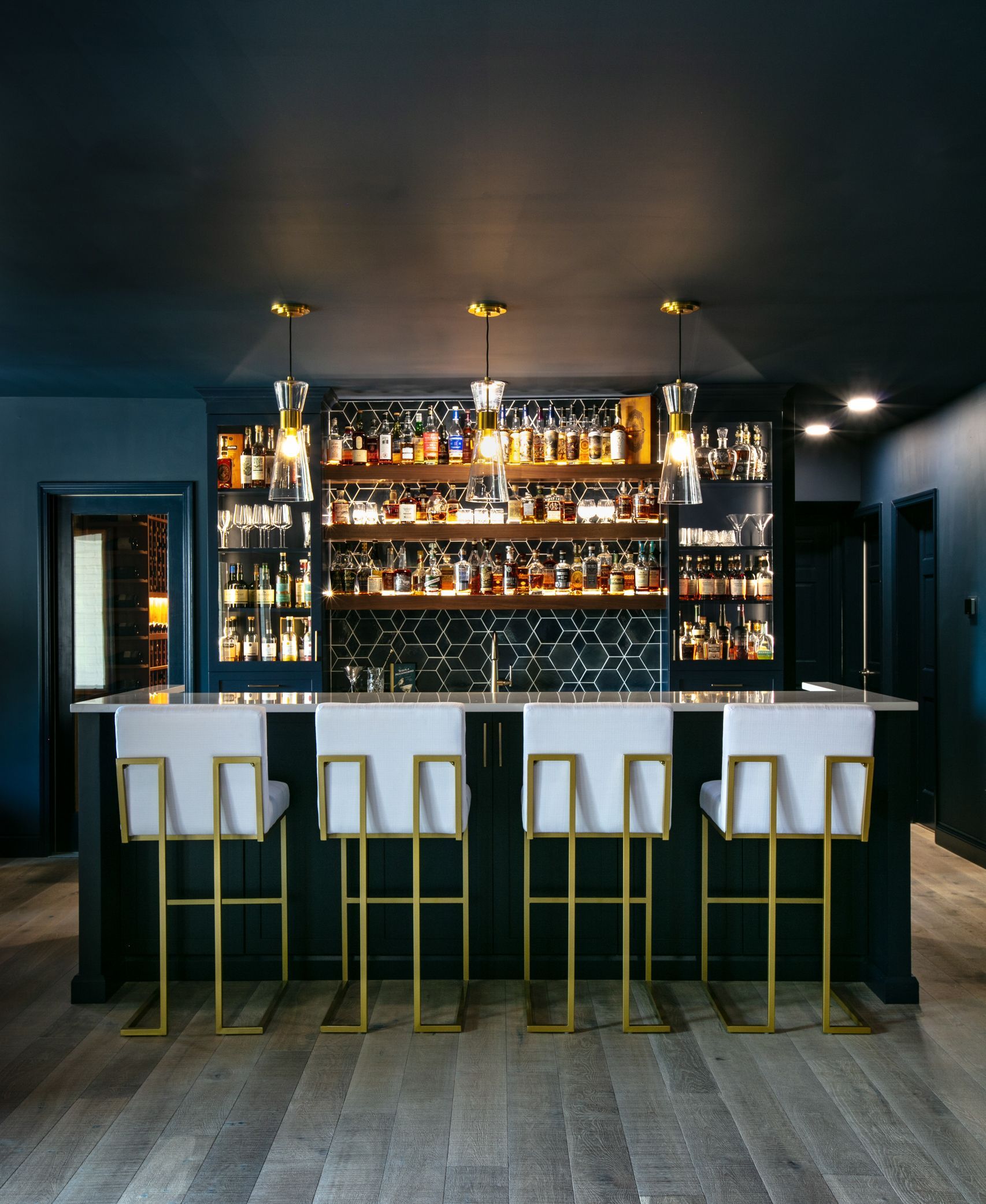 Dark home bar with four white and gold bar stools and shelves filled with bottles.