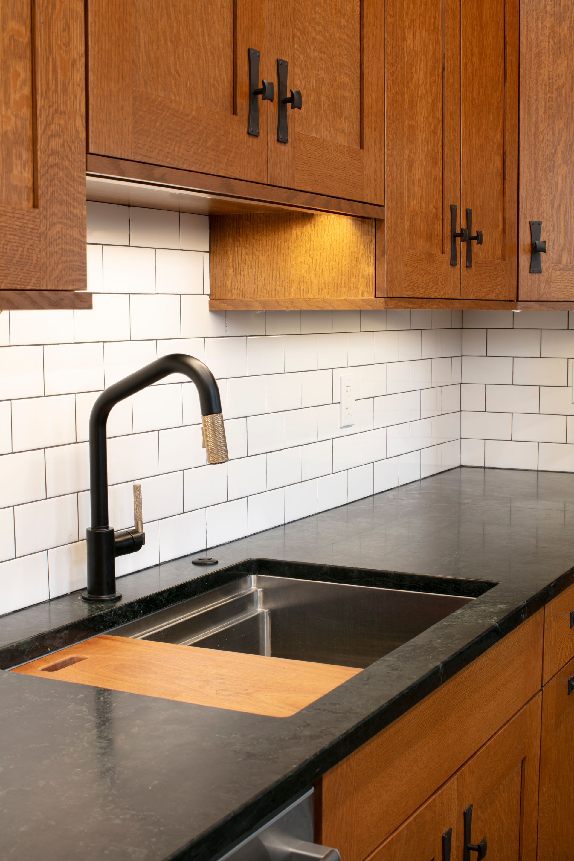 Kitchen with wooden cabinets, black countertop, stainless steel sink, subway tile backsplash, and black faucet.
