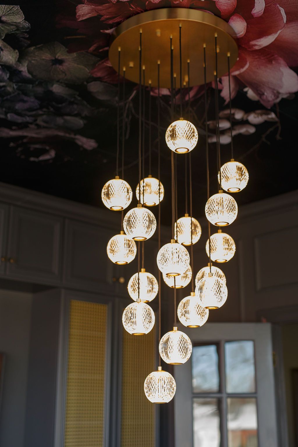 Gold chandelier with hanging globes illuminating a room with floral ceiling wallpaper.