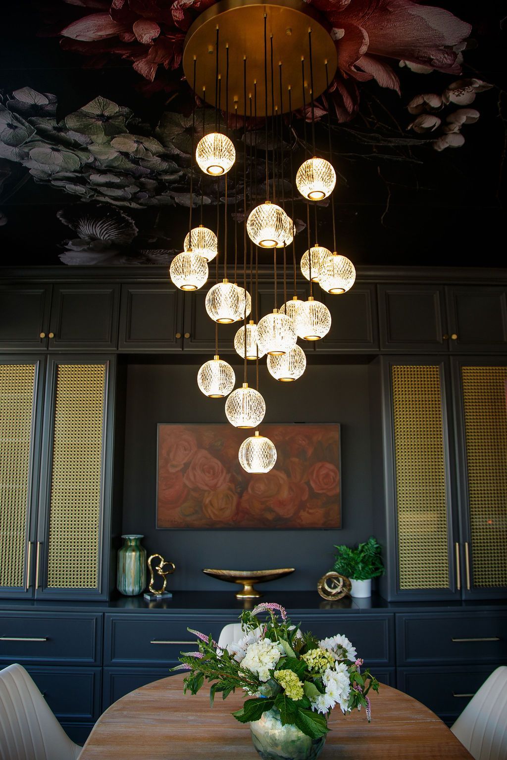Dining room with dark cabinets, floral ceiling, and a large chandelier over an oval table with a floral centerpiece.
