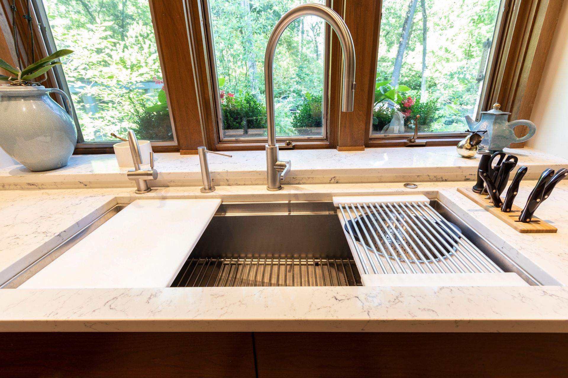 Stainless steel kitchen sink with accessories set against a window with a lush green view.