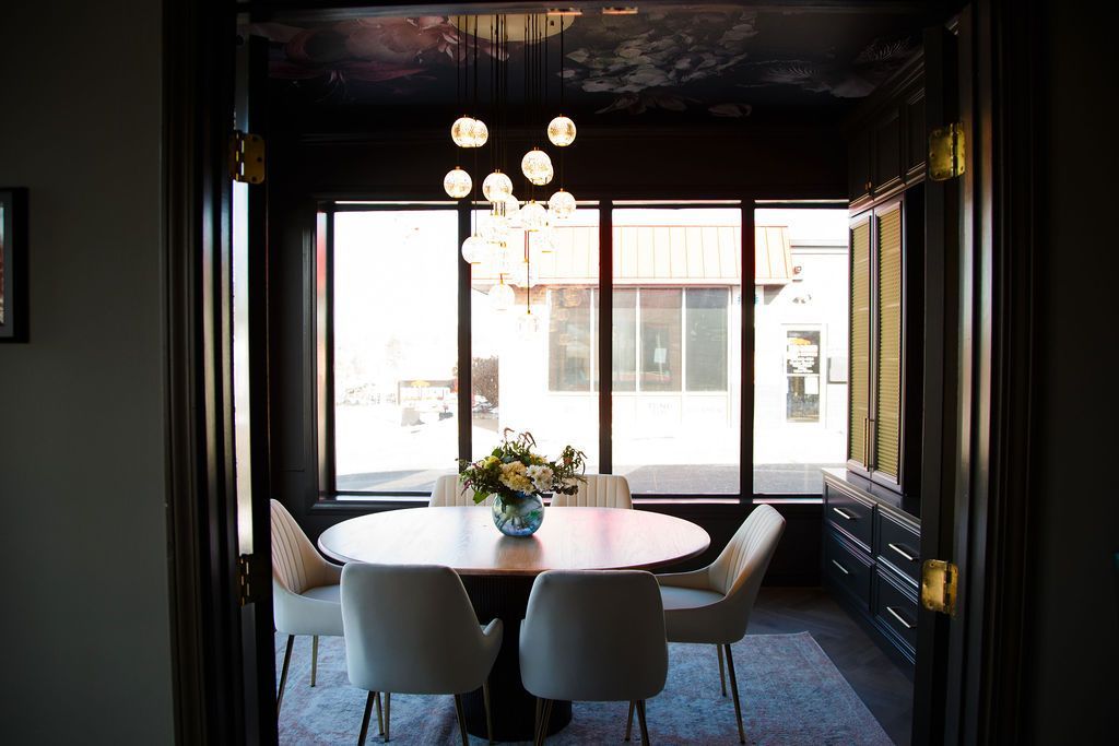 Dining room with round table, chairs, chandelier, and a large window overlooking a street.