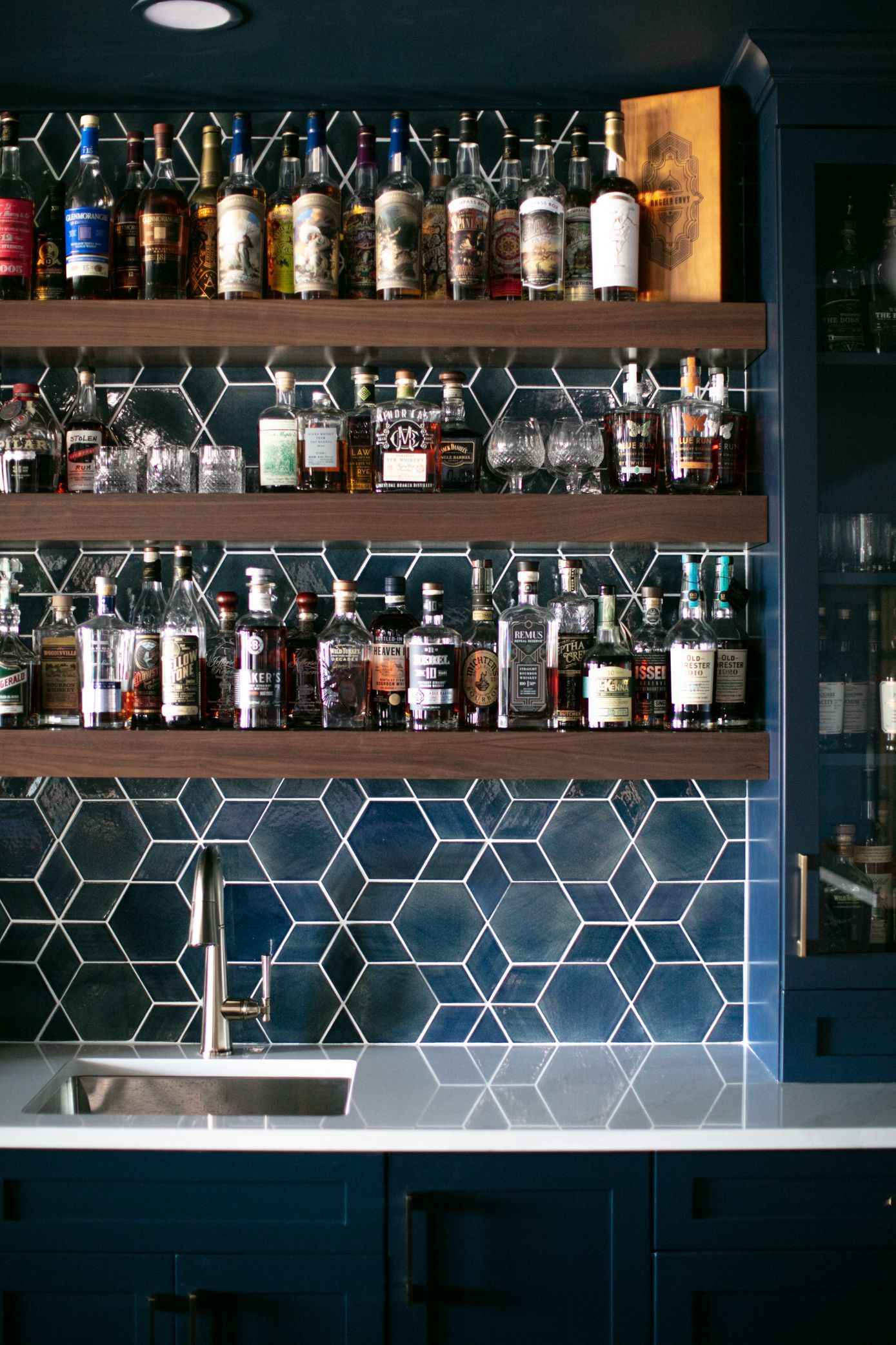 Home bar with dark blue cabinets, shelves of liquor, and geometric tile backsplash.