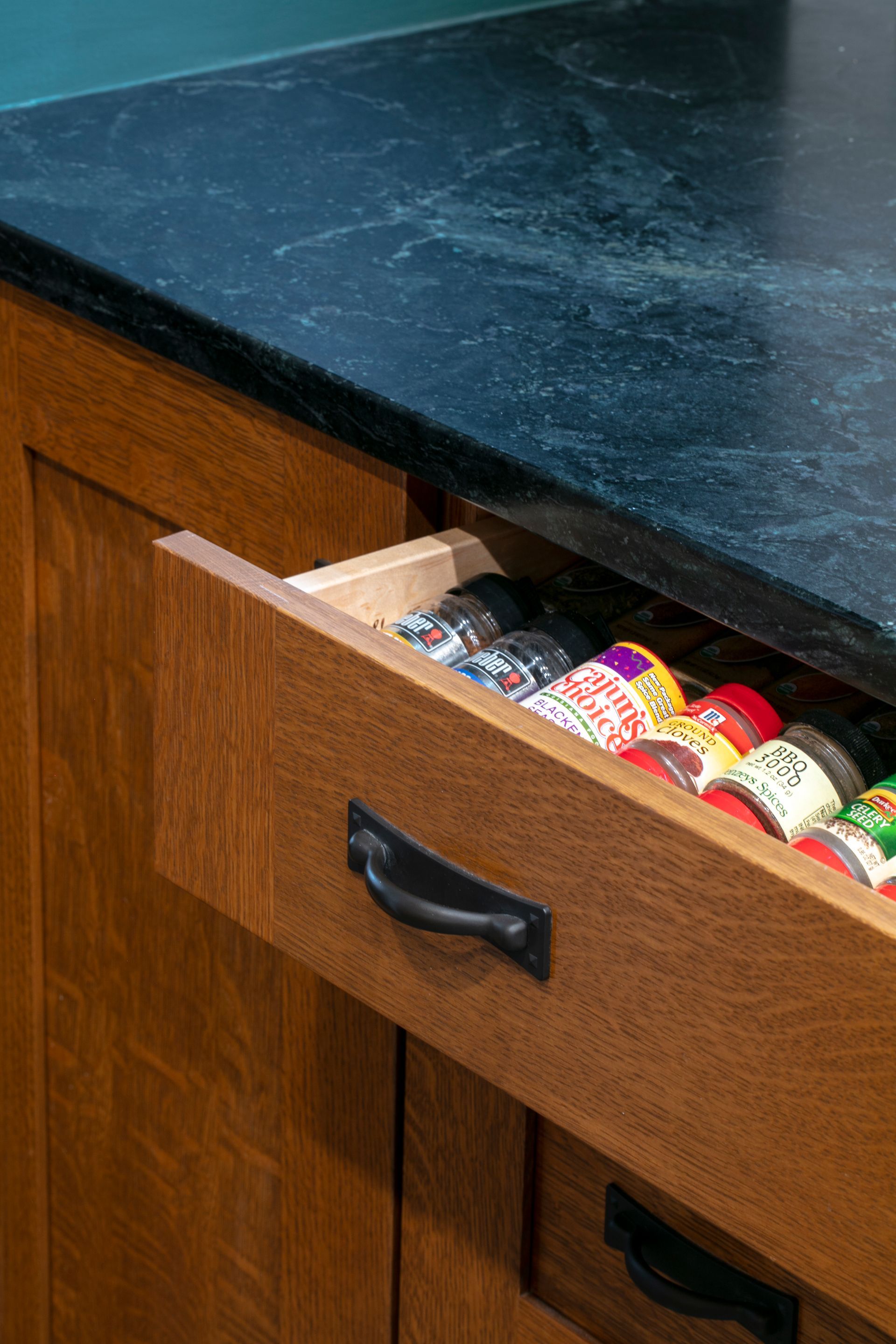 Open kitchen drawer with spice bottles. Black countertop and wooden cabinets.