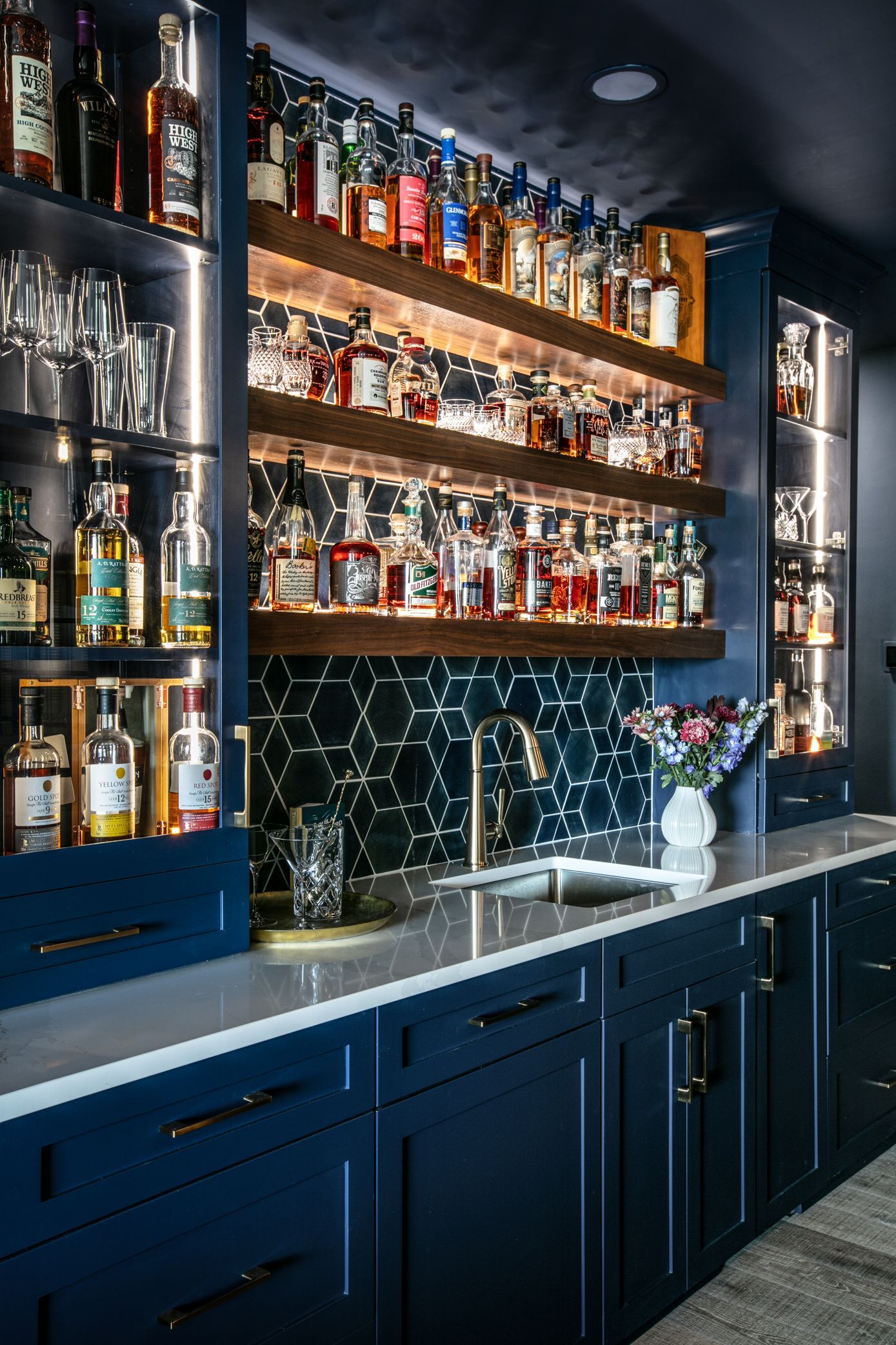 A home bar with dark blue cabinetry, shelves filled with liquor bottles, and a patterned backsplash.