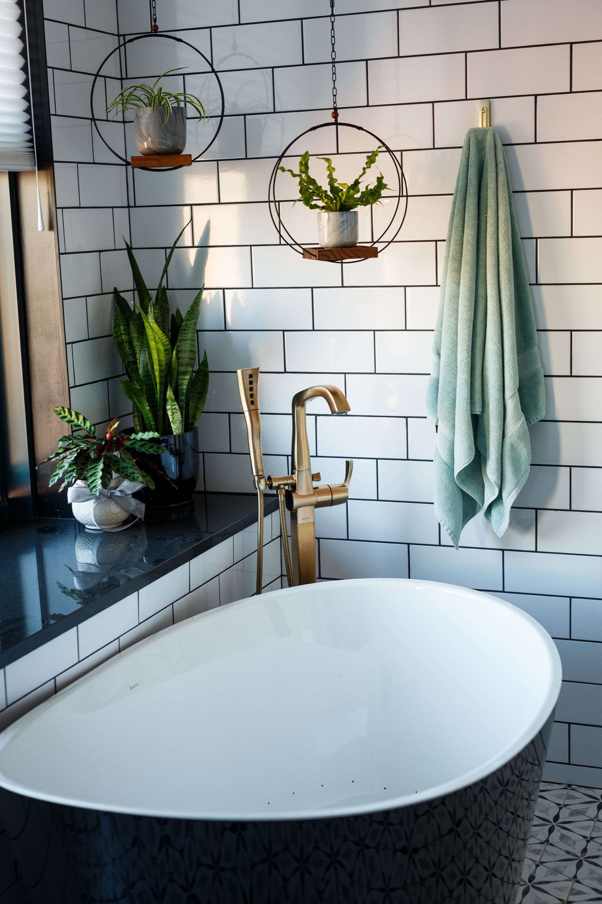 A modern bathroom with a black and white tiled wall, a gold faucet, a tub, and hanging plants.