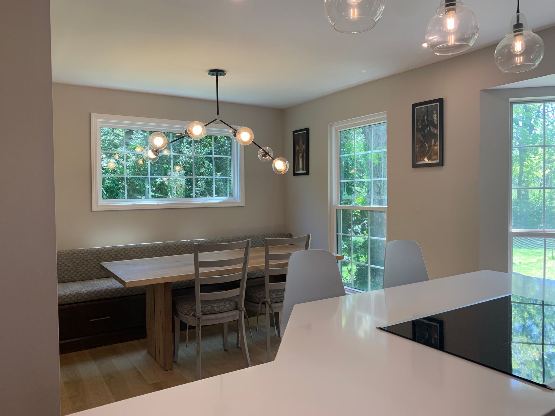 Dining room with a table, built-in bench, and windows overlooking greenery. Neutral-toned walls and modern light fixtures.