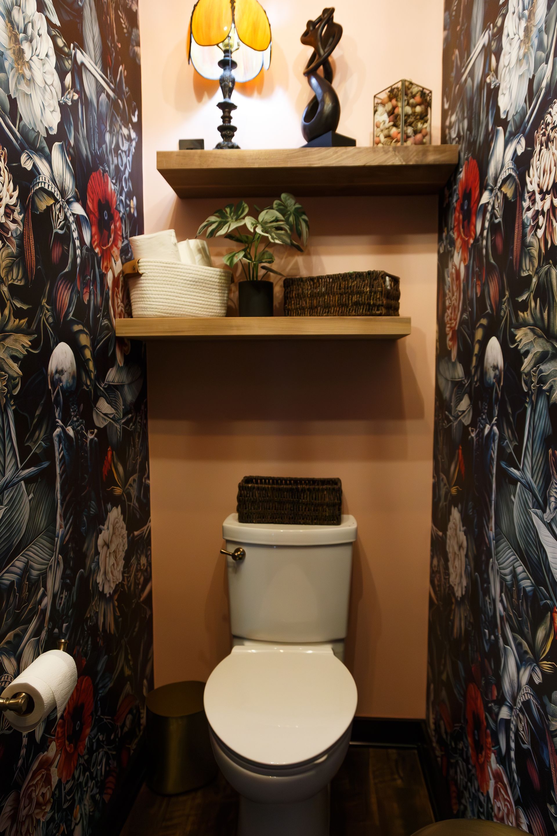 Small bathroom with floral wallpaper, pink walls, wood shelves with decor, and a white toilet.