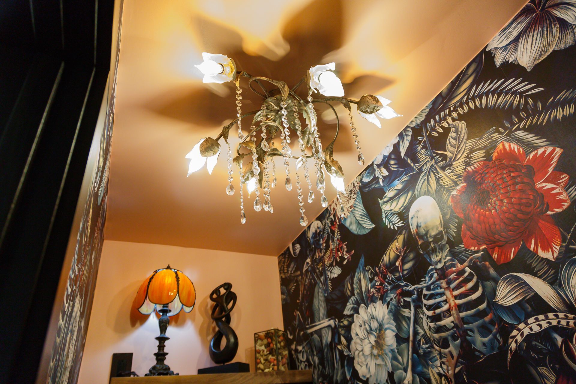Ornate chandelier hanging on a pink ceiling, with floral wallpaper and a lamp below.