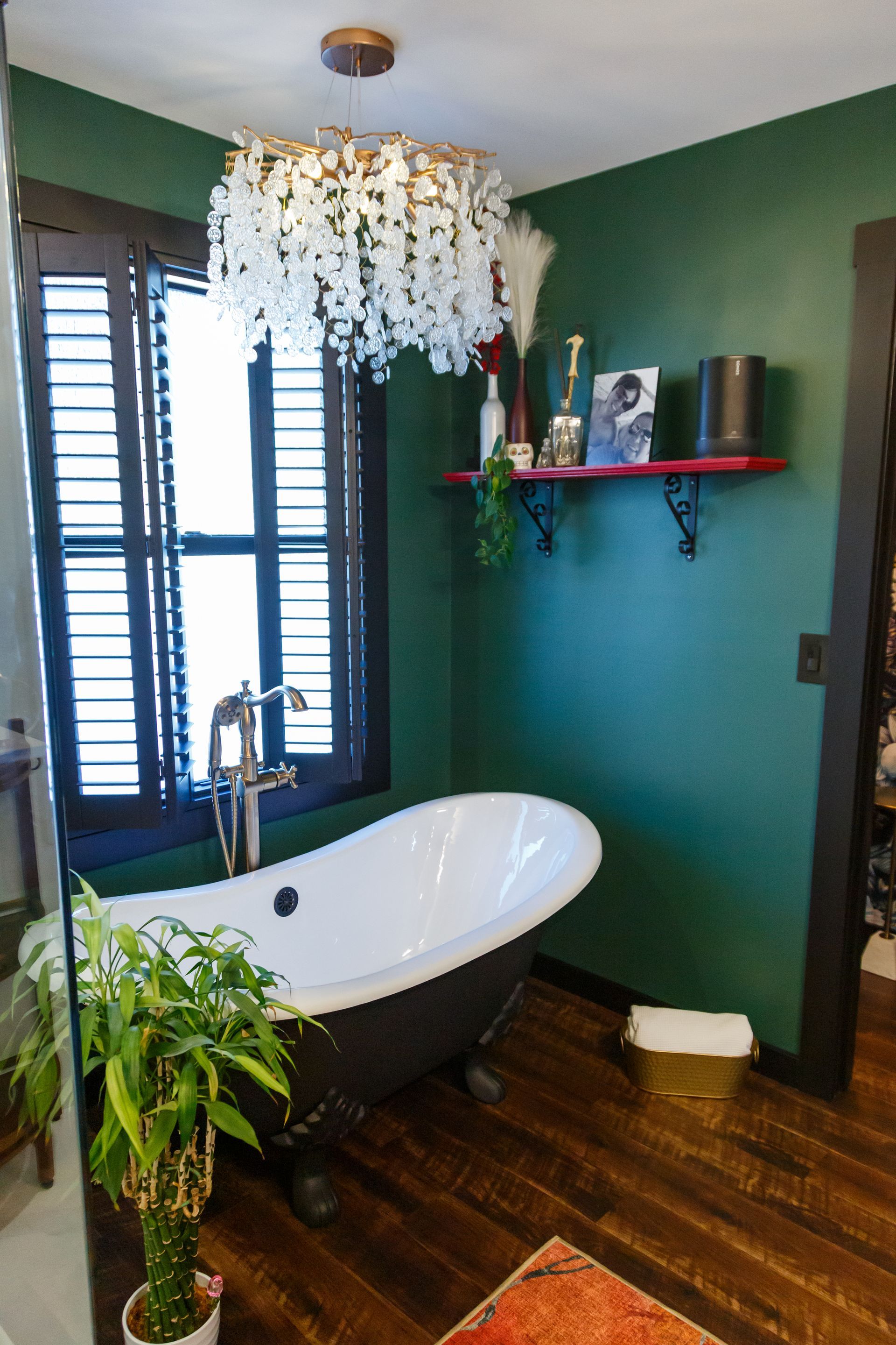 Bathroom with clawfoot tub, dark green walls, wood shutters, and ornate chandelier.