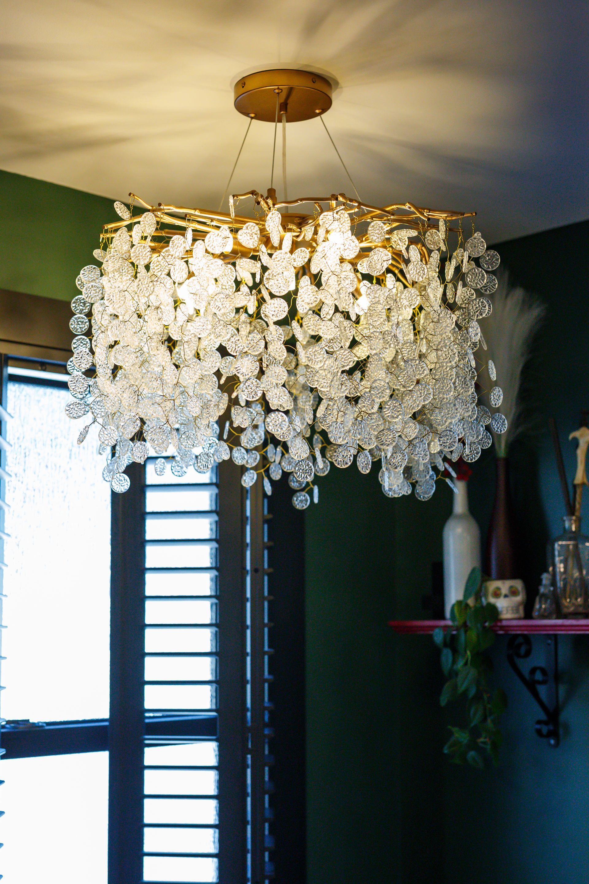 Gold chandelier with hanging crystal droplets, in a room with a window and green walls.