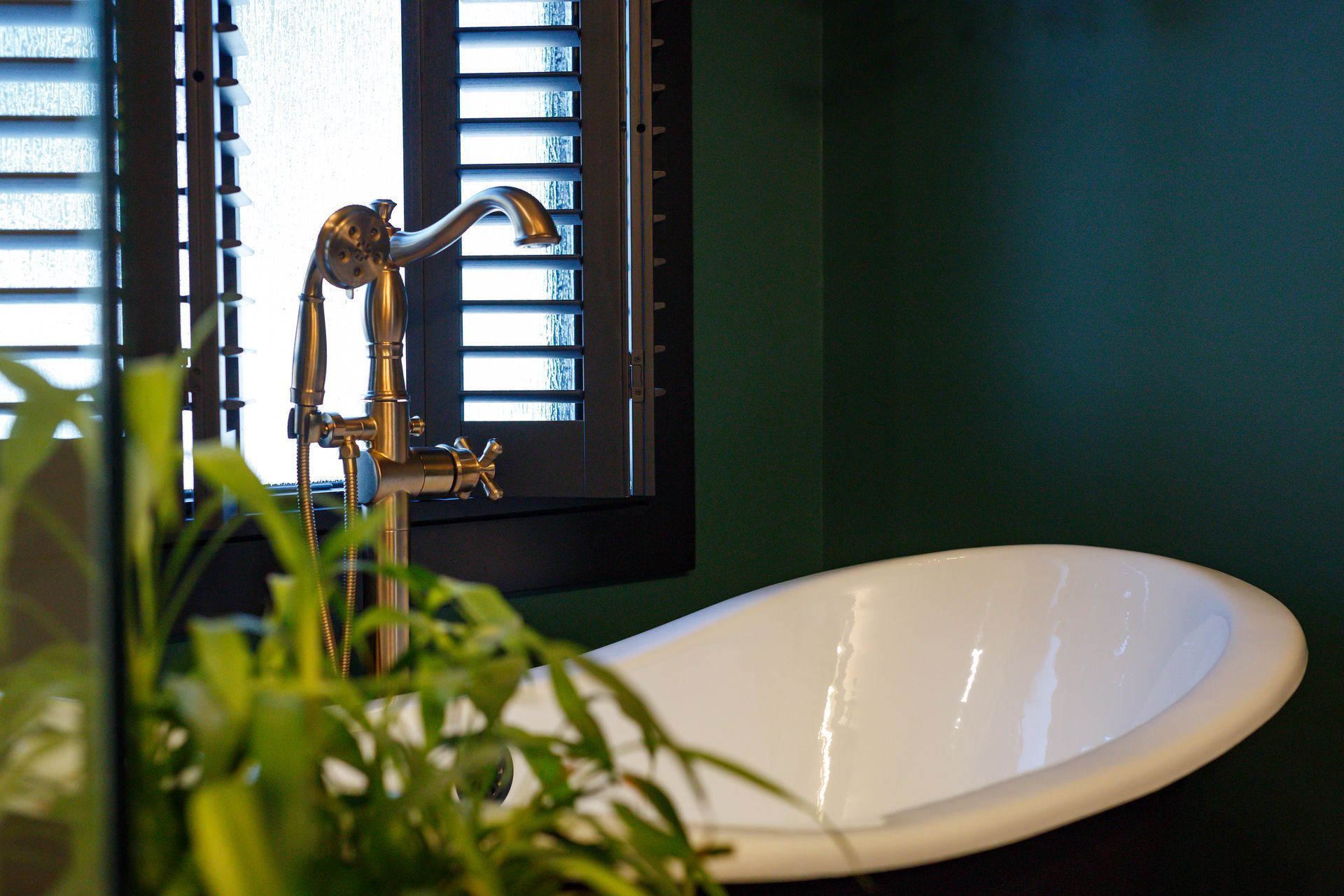 Claw-foot bathtub with gold faucet near a window with shutters. Dark green wall. A plant in the foreground.