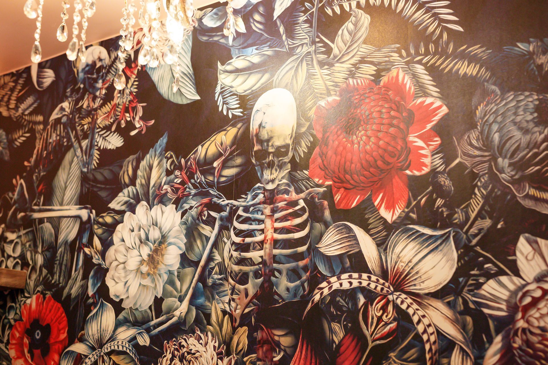 Skeleton in floral wallpaper, large red flower prominent. Dark background, chandelier visible.