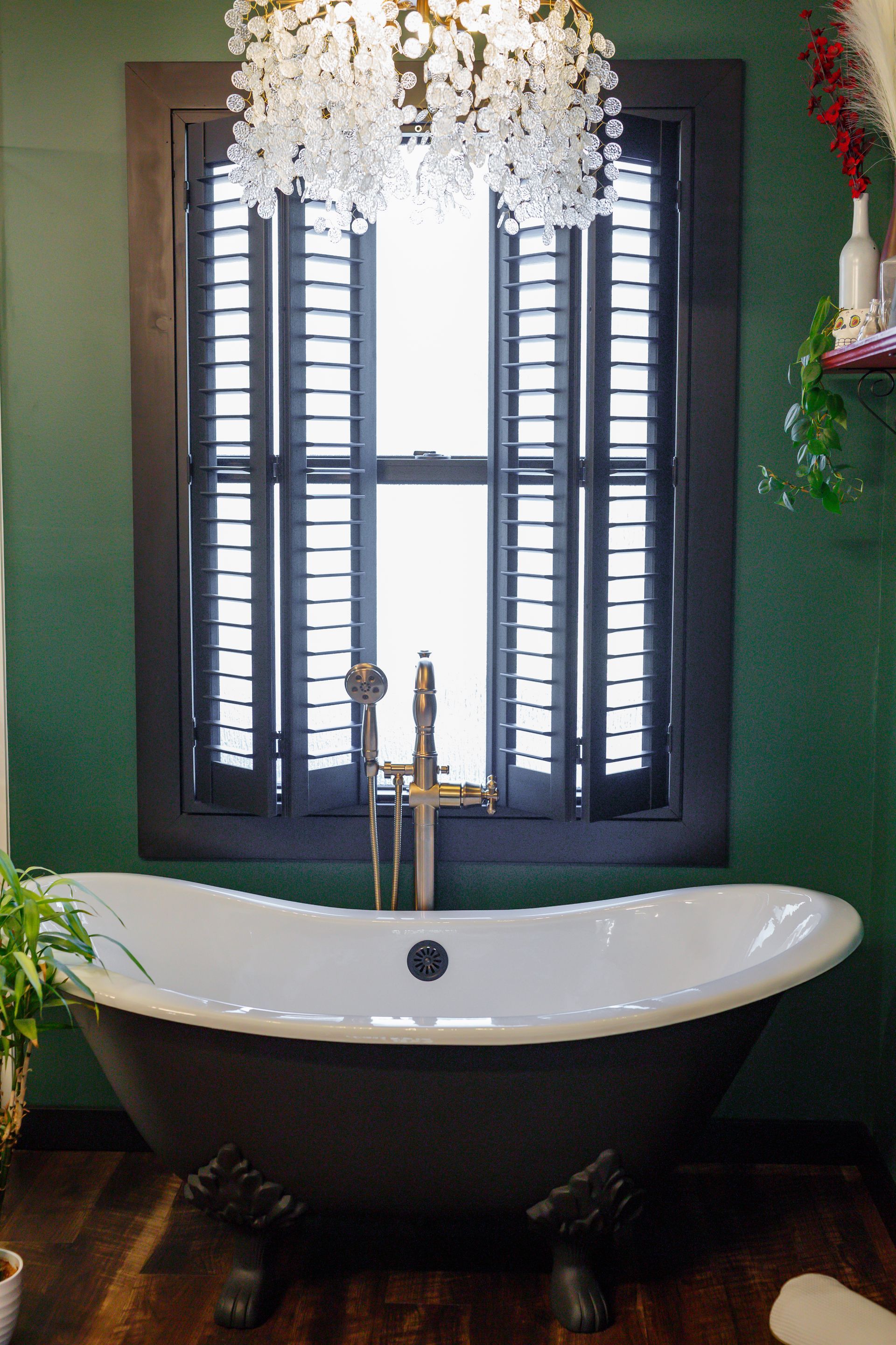 Clawfoot bathtub under a window with black shutters, in a green-walled bathroom.