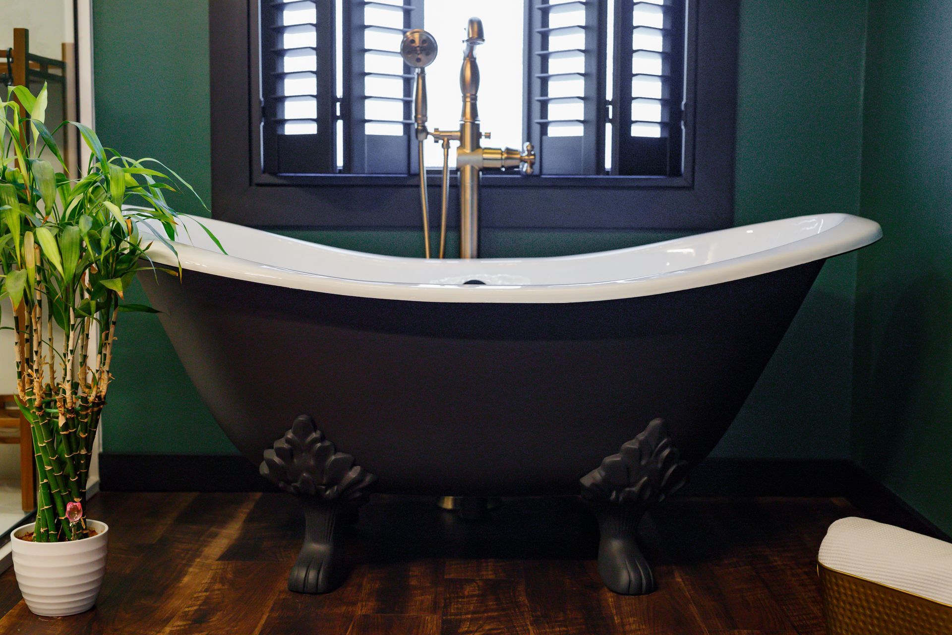 Claw-footed black bathtub with brass fixtures, in front of a window with shutters.