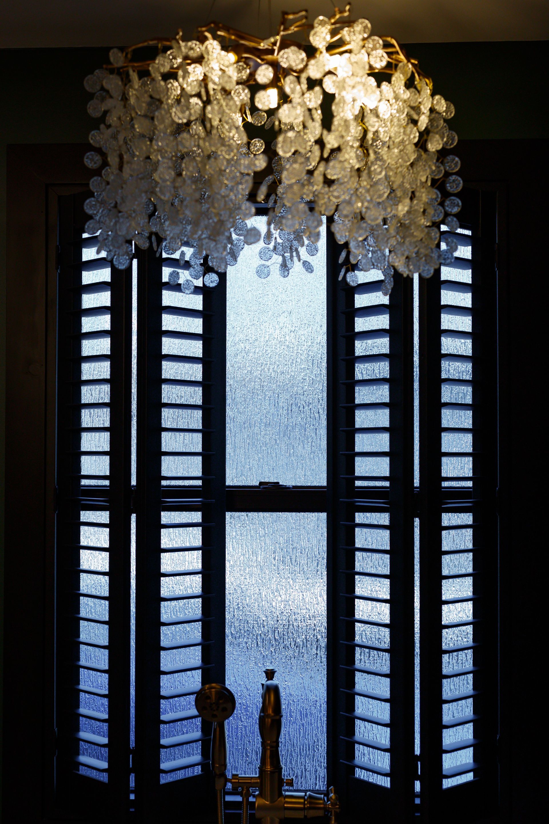 Chandelier above a window with open shutters; light shines through textured glass.