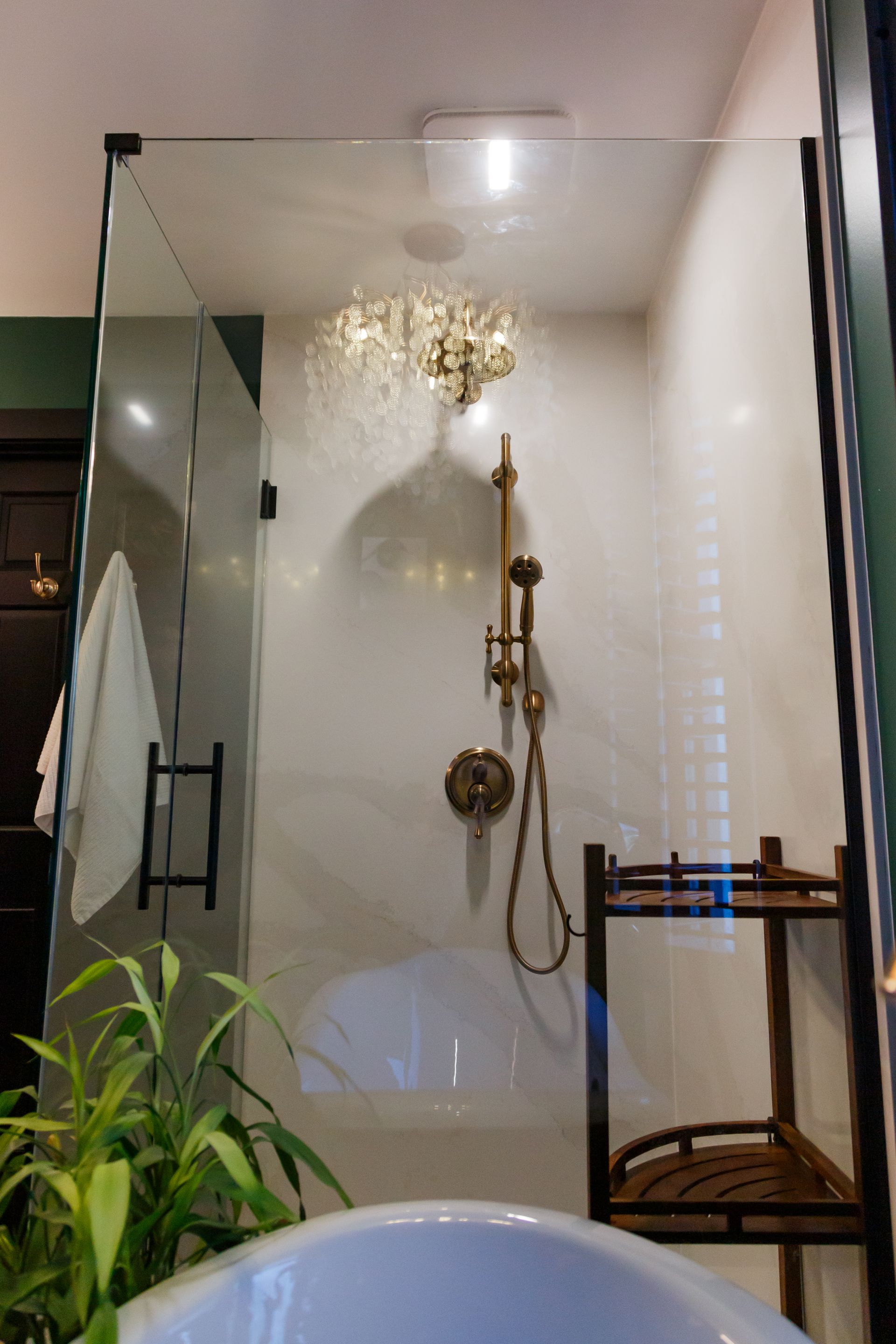 Glass shower with gold fixtures and a chandelier, next to a black shelf and green plant.