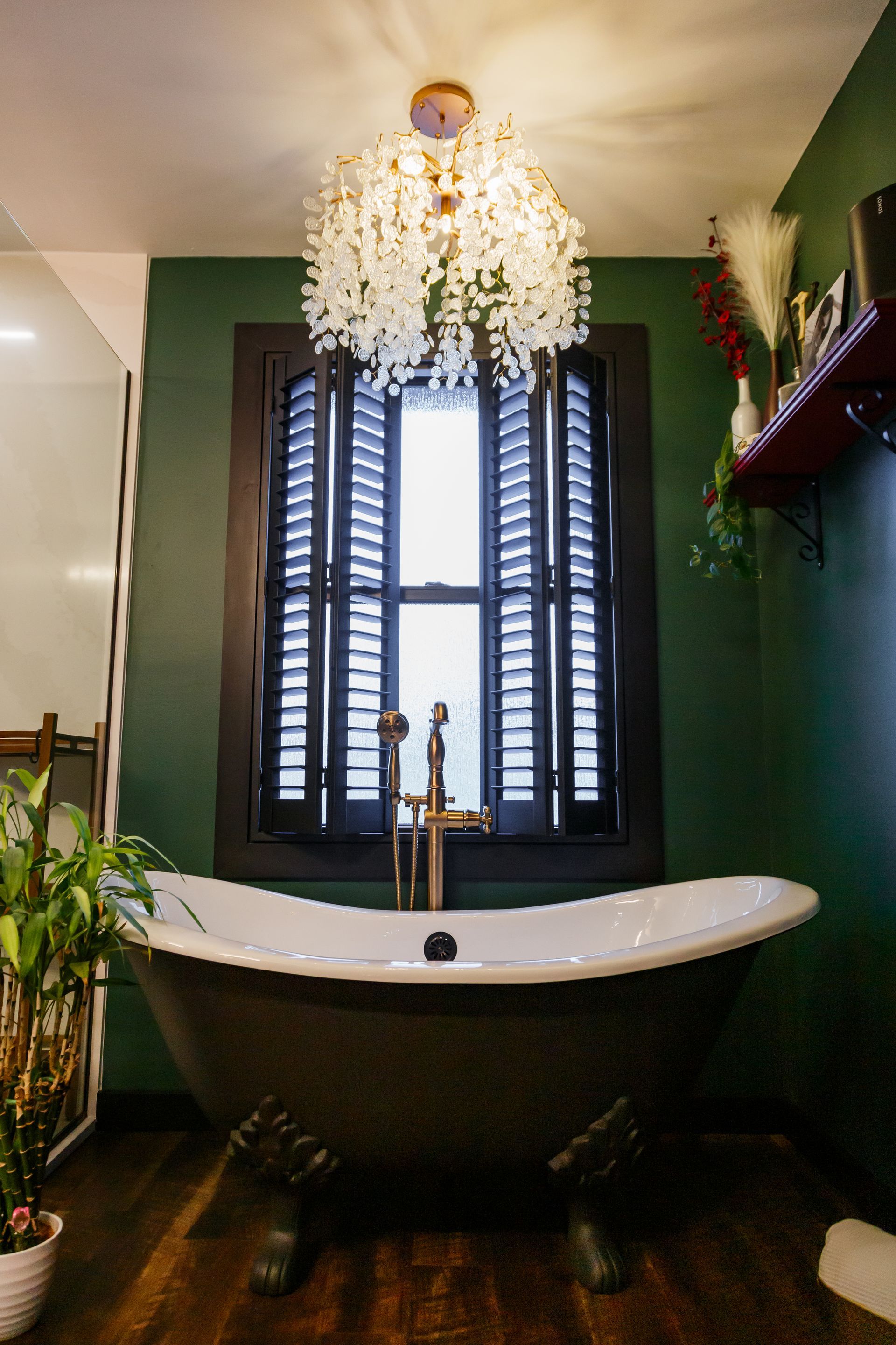 Claw-foot bathtub under a window with shutters. Dark green walls, gold faucet, and a decorative light fixture.