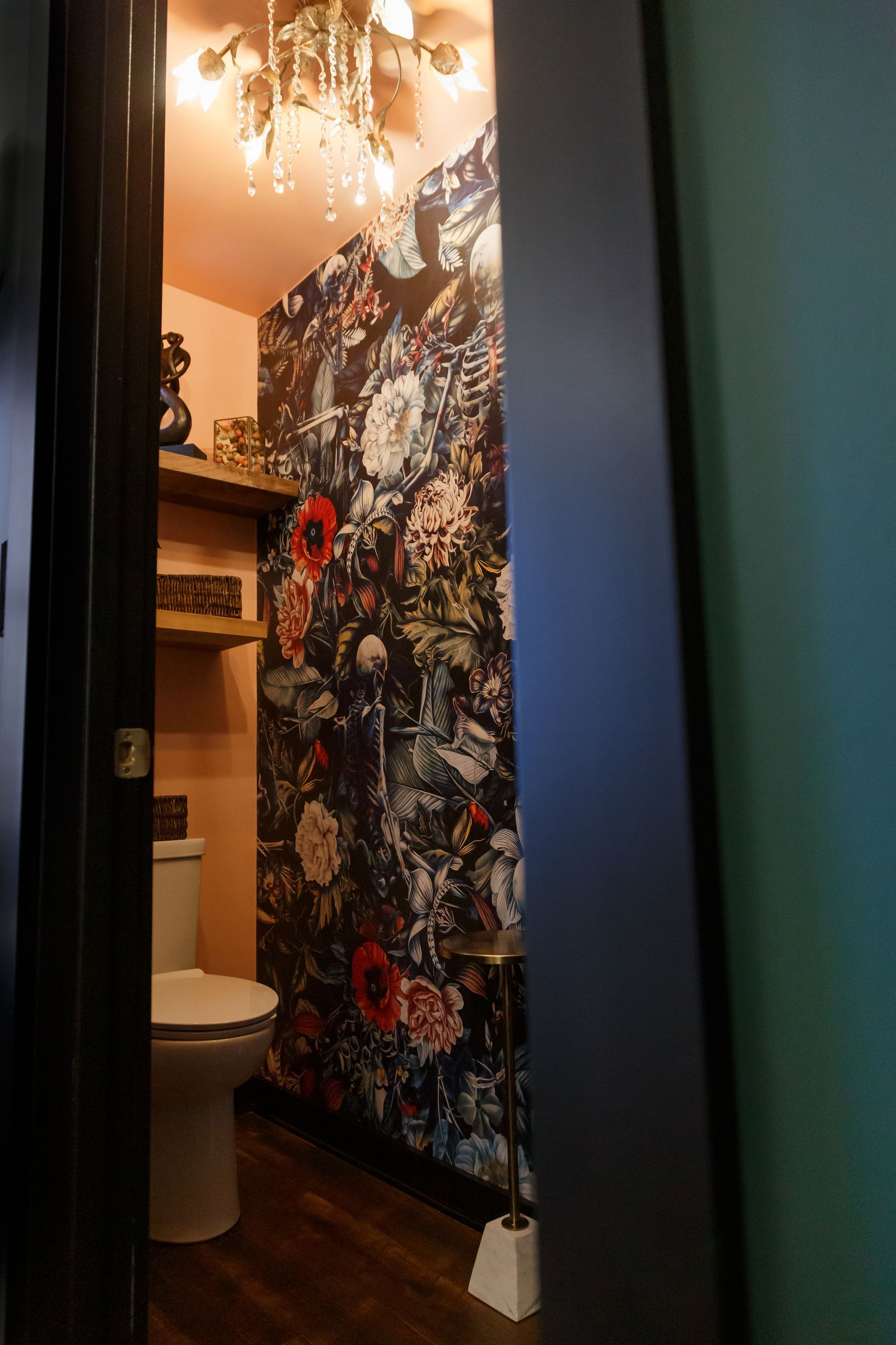 A small powder room with floral wallpaper and a gold chandelier. A toilet, shelves, and a toilet brush are visible.