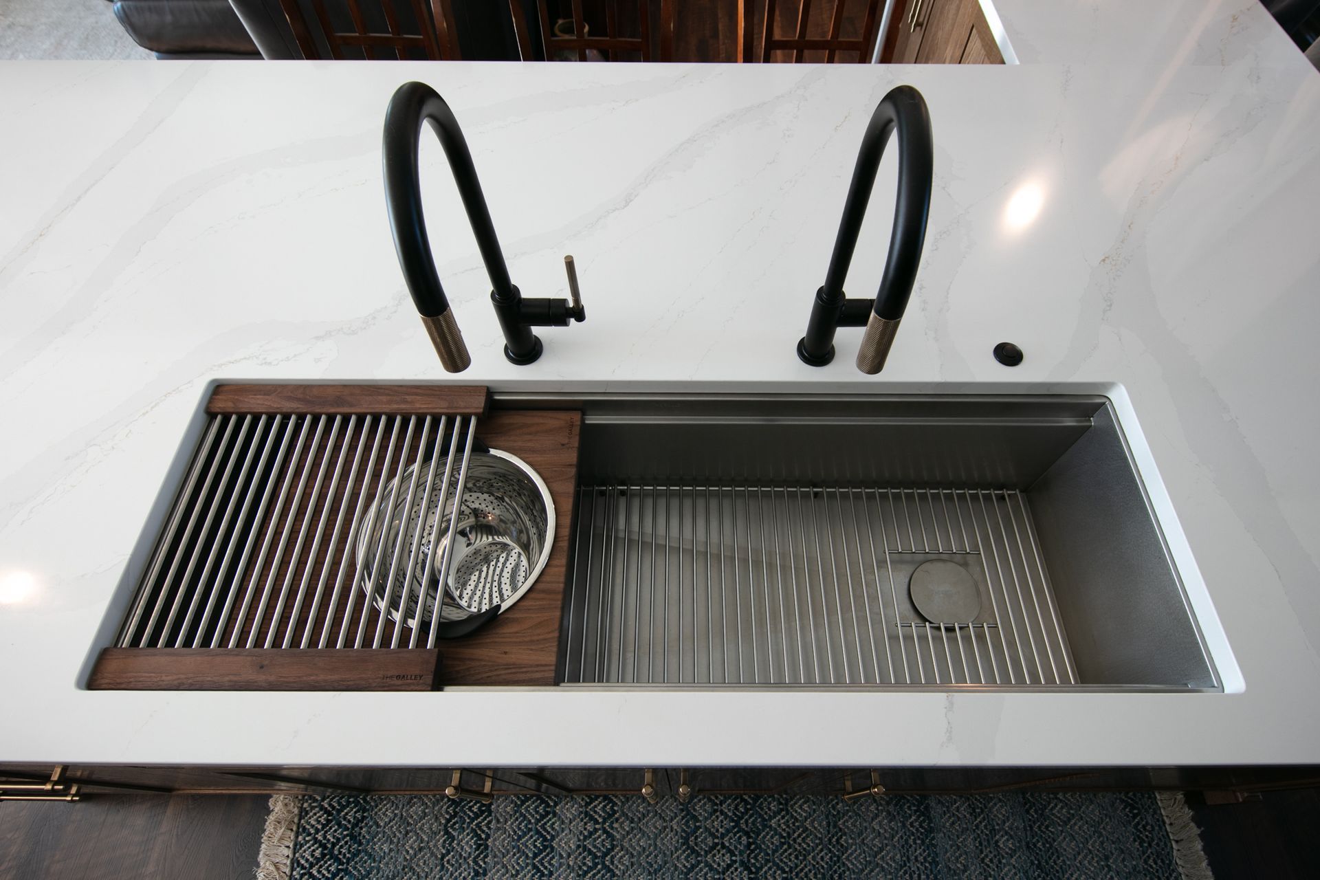Overhead view of a modern kitchen sink with a stainless steel basin, wooden accessories, and two black faucets on a white countertop.