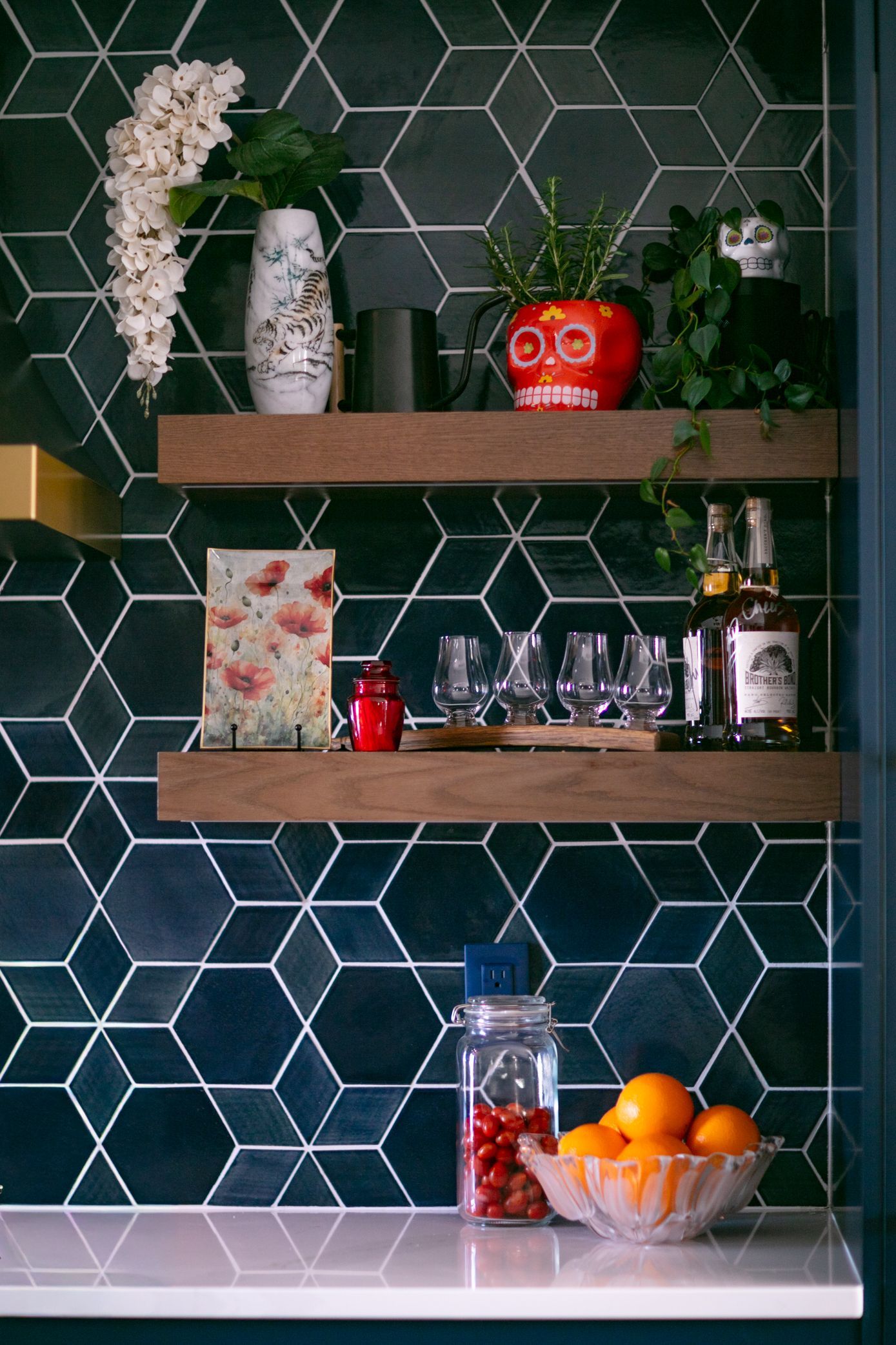 Shelves with barware and decor against a dark hexagon-tiled wall. Includes glasses, liquor, and a red skull.