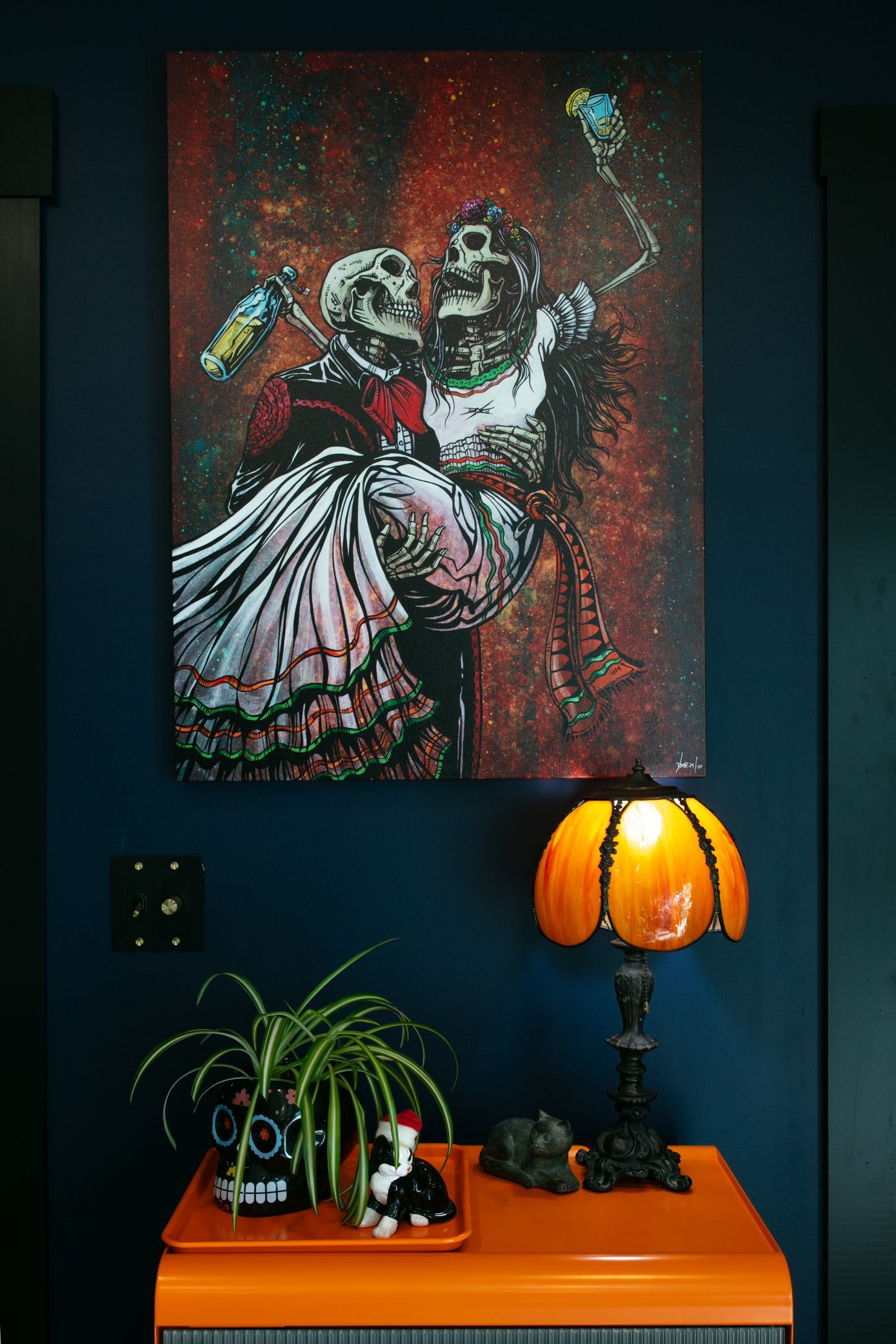 Skeletal figures dancing on a canvas above an orange table, with a pumpkin lamp, plant, and decor against a dark blue wall.