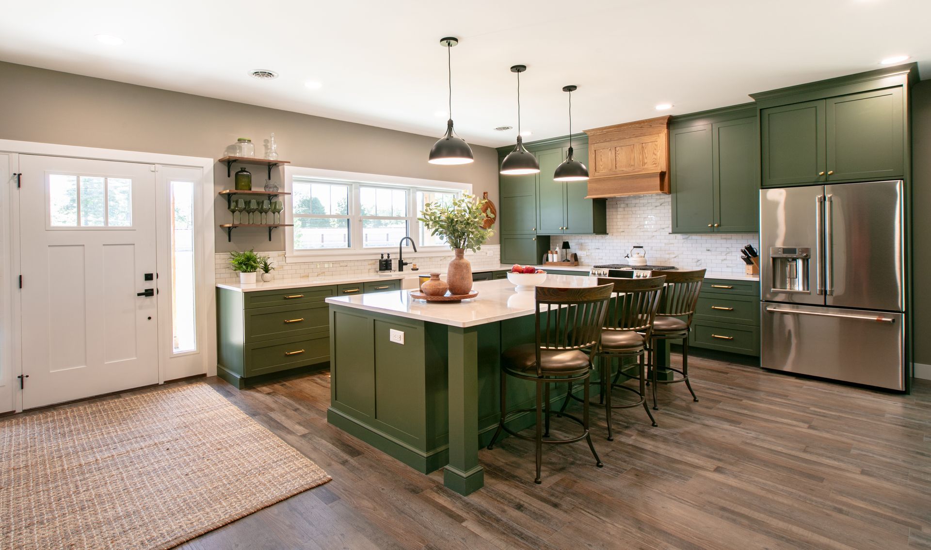 Green kitchen with island, stainless steel refrigerator, and wooden floors.