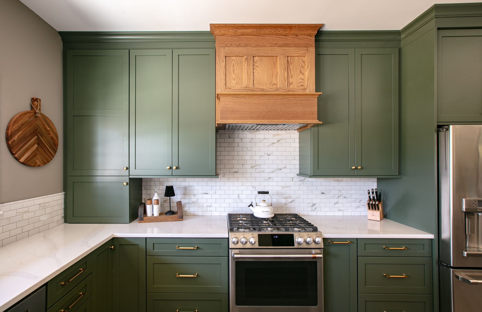 Green kitchen cabinets with white countertops, wooden range hood, and stainless steel appliances.