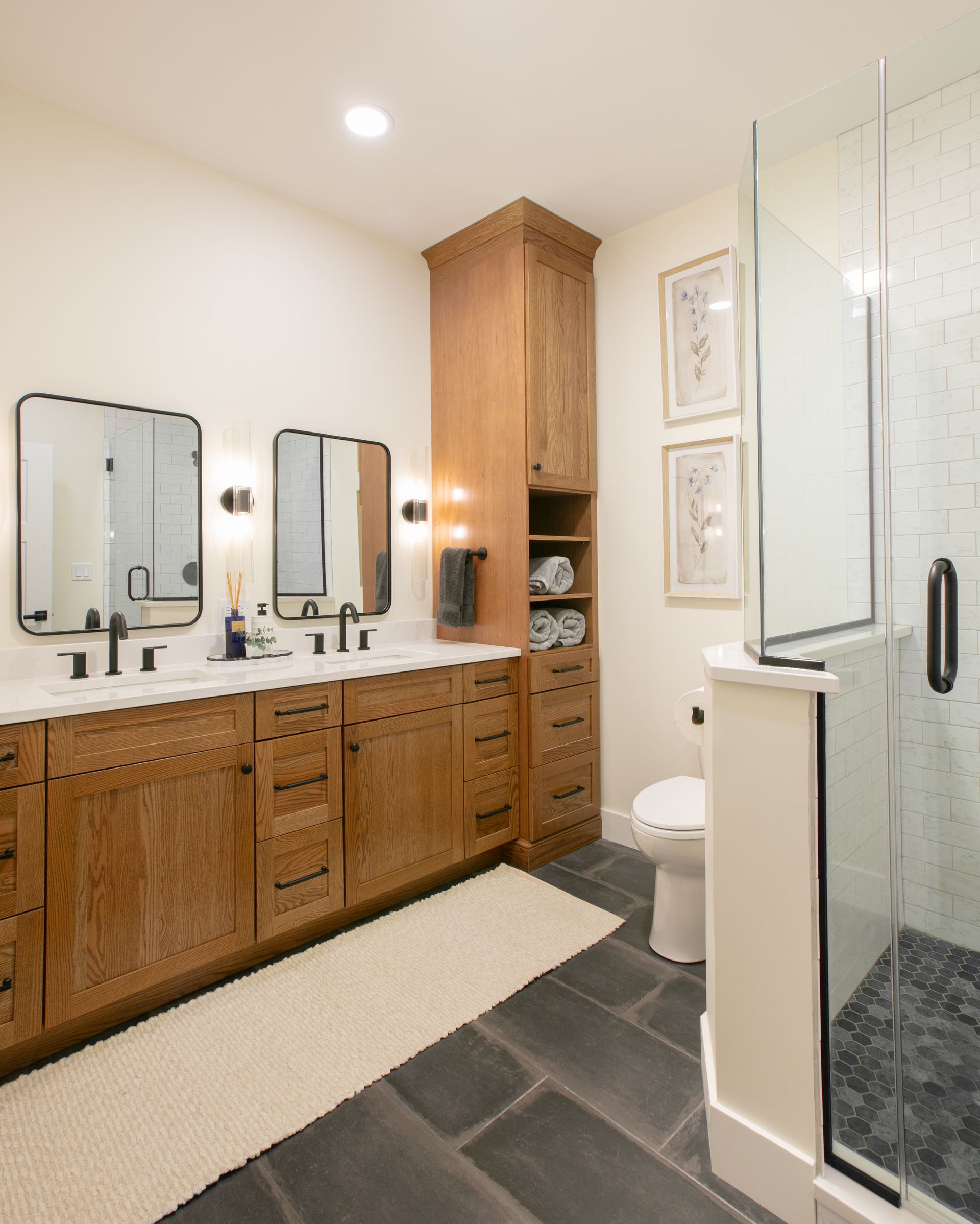 Bathroom with wooden cabinets, dual sinks, mirrors, shower, and dark tile floor.