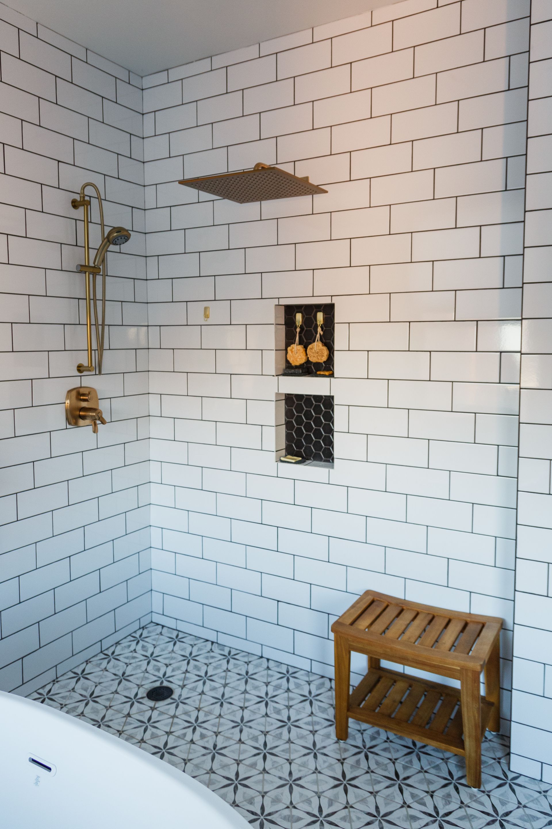 A modern white-tiled shower with gold fixtures, built-in shelving, a wooden bench, and patterned floor.