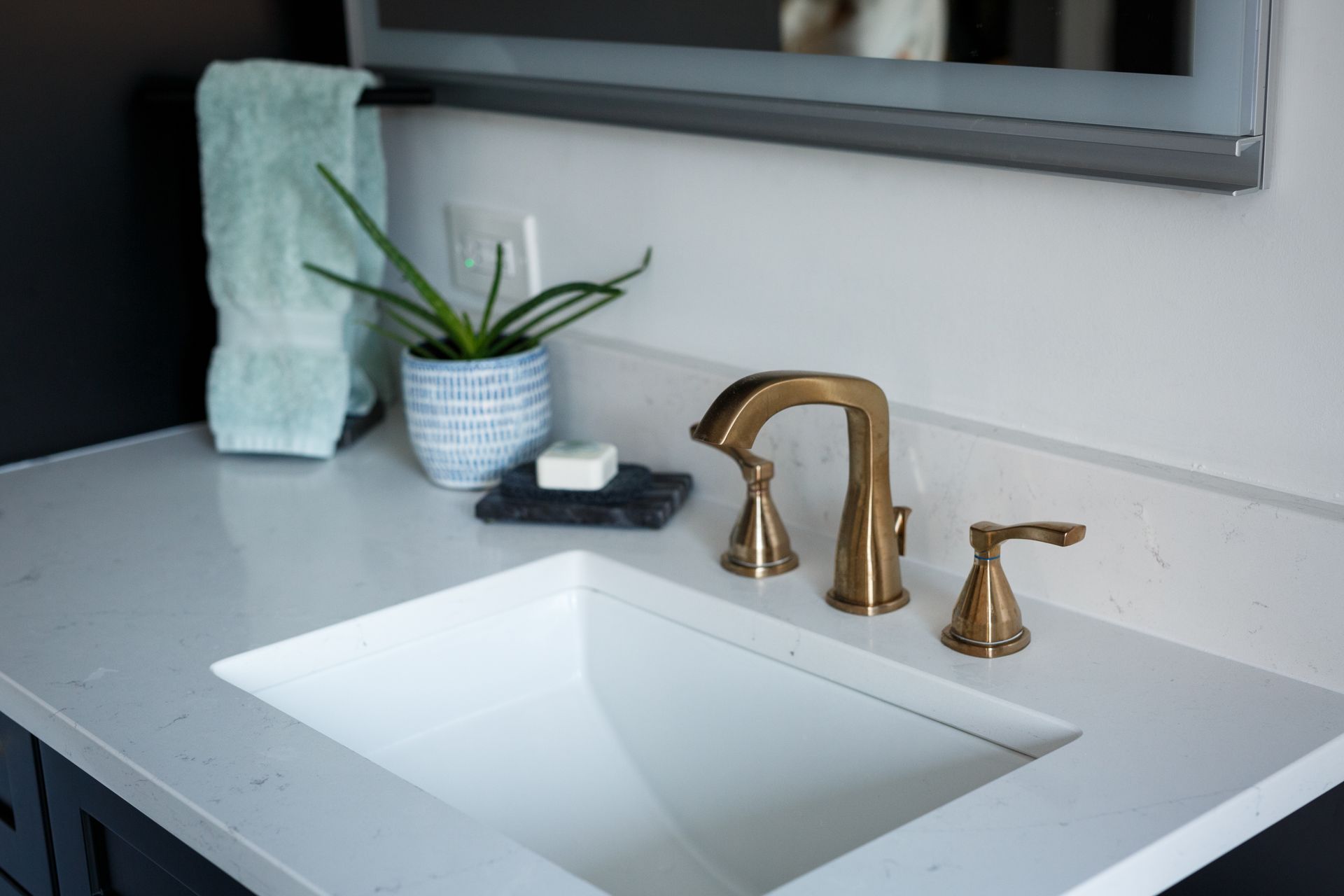 Bathroom sink with gold faucet, soap, plant, and towel. White countertop, black cabinet, and mirror.