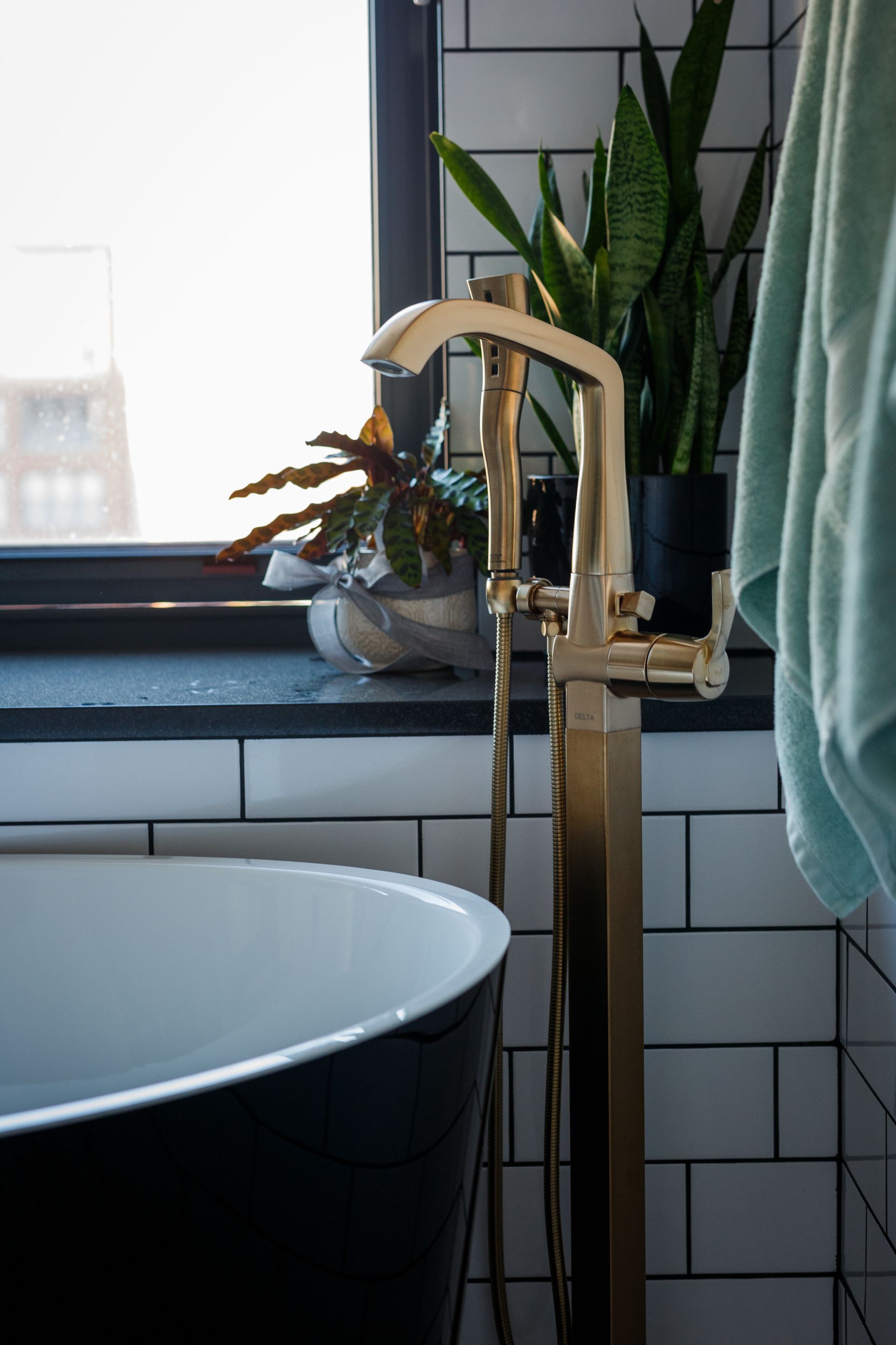 Black bathtub, gold faucet, and potted plants next to a window in a bathroom with white subway tile.