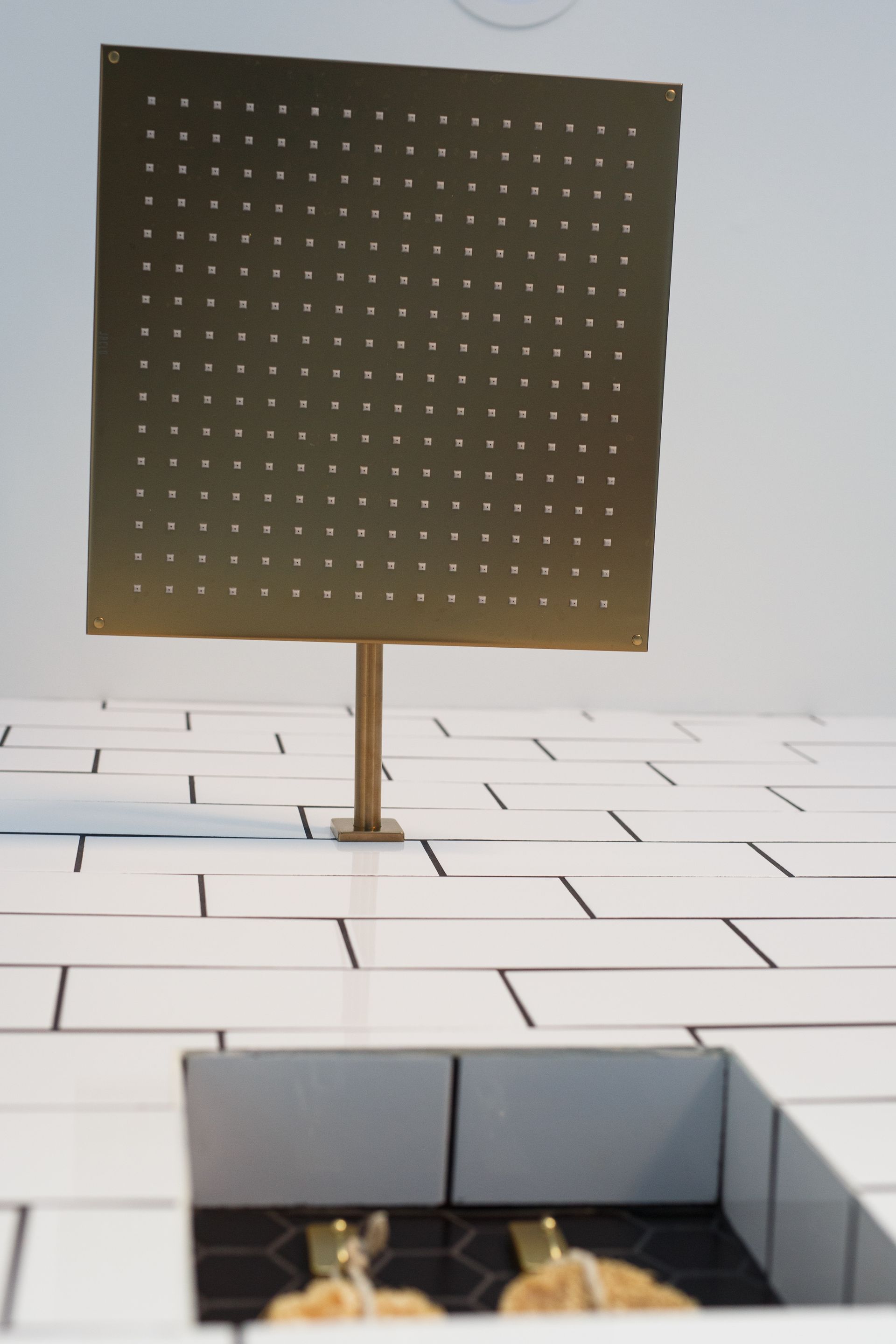 Square gold shower head with evenly spaced holes, over white tiled wall and black tile inset.